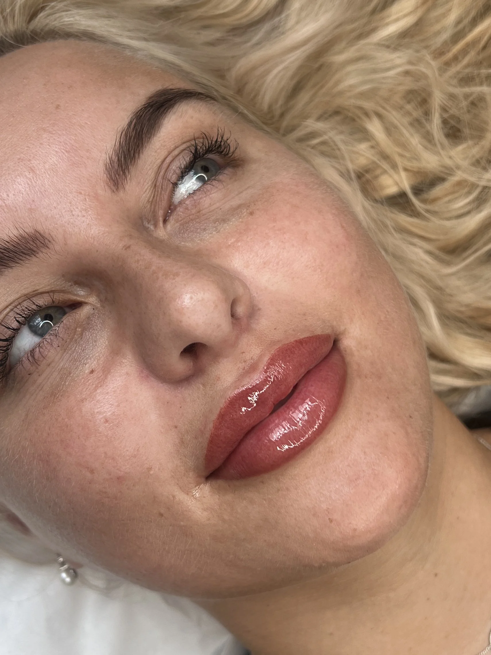 Close-up of a woman's face with light makeup, blonde curly hair, and blue eyes, lying down and looking upward. she's had her lips tattooed
