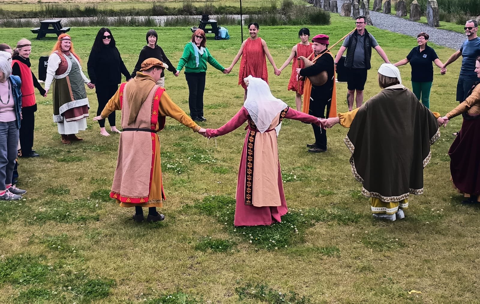 A group of people holding hands in a circle on a grassy field, some wearing historical costumes, participating in a communal activity or dance.
