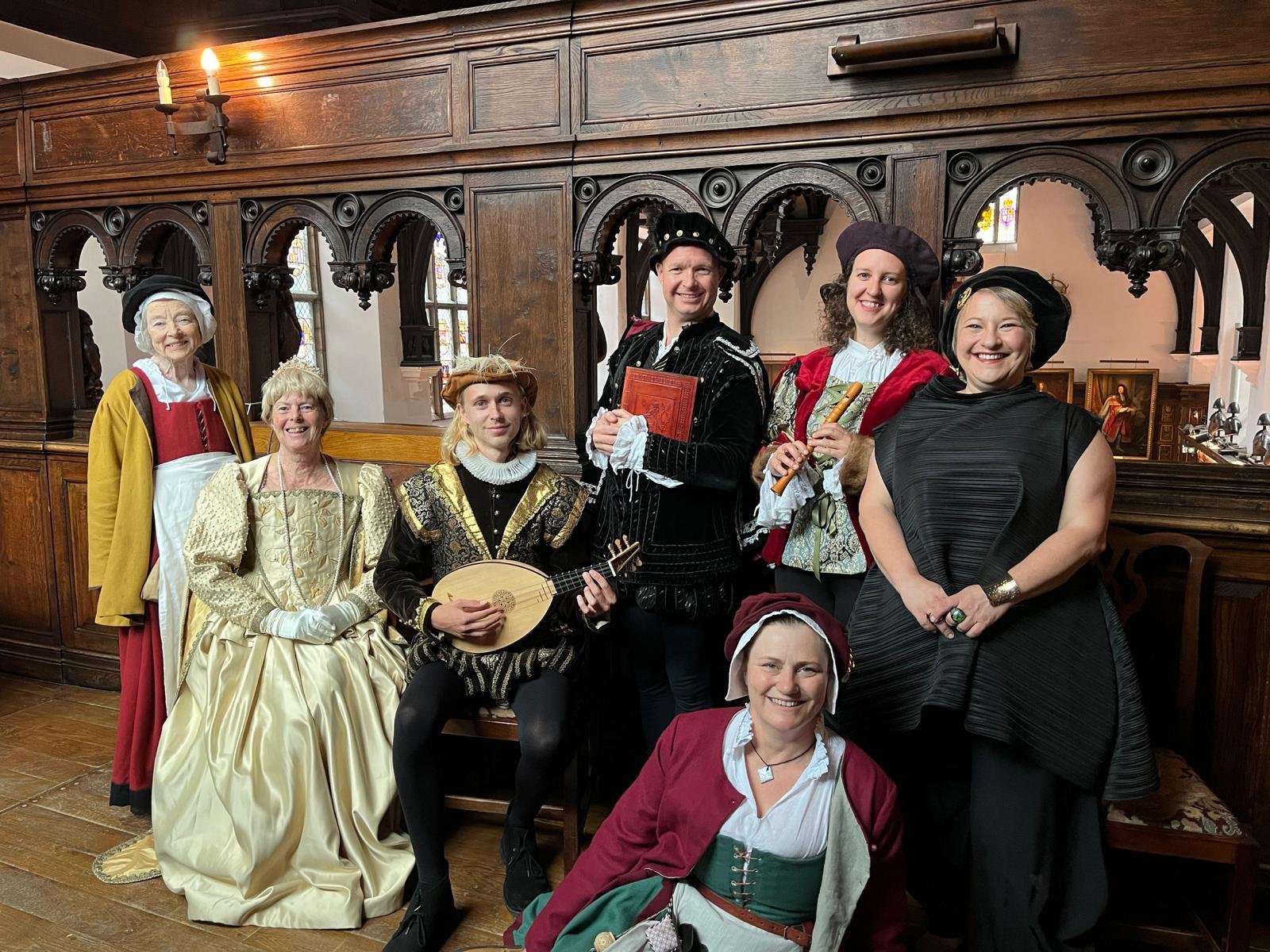 A group of people dressed in Renaissance-era costumes, posing in a historic room with wooden paneling. Some wear dresses and others wear doublets, with accessories like hats and musical instruments.