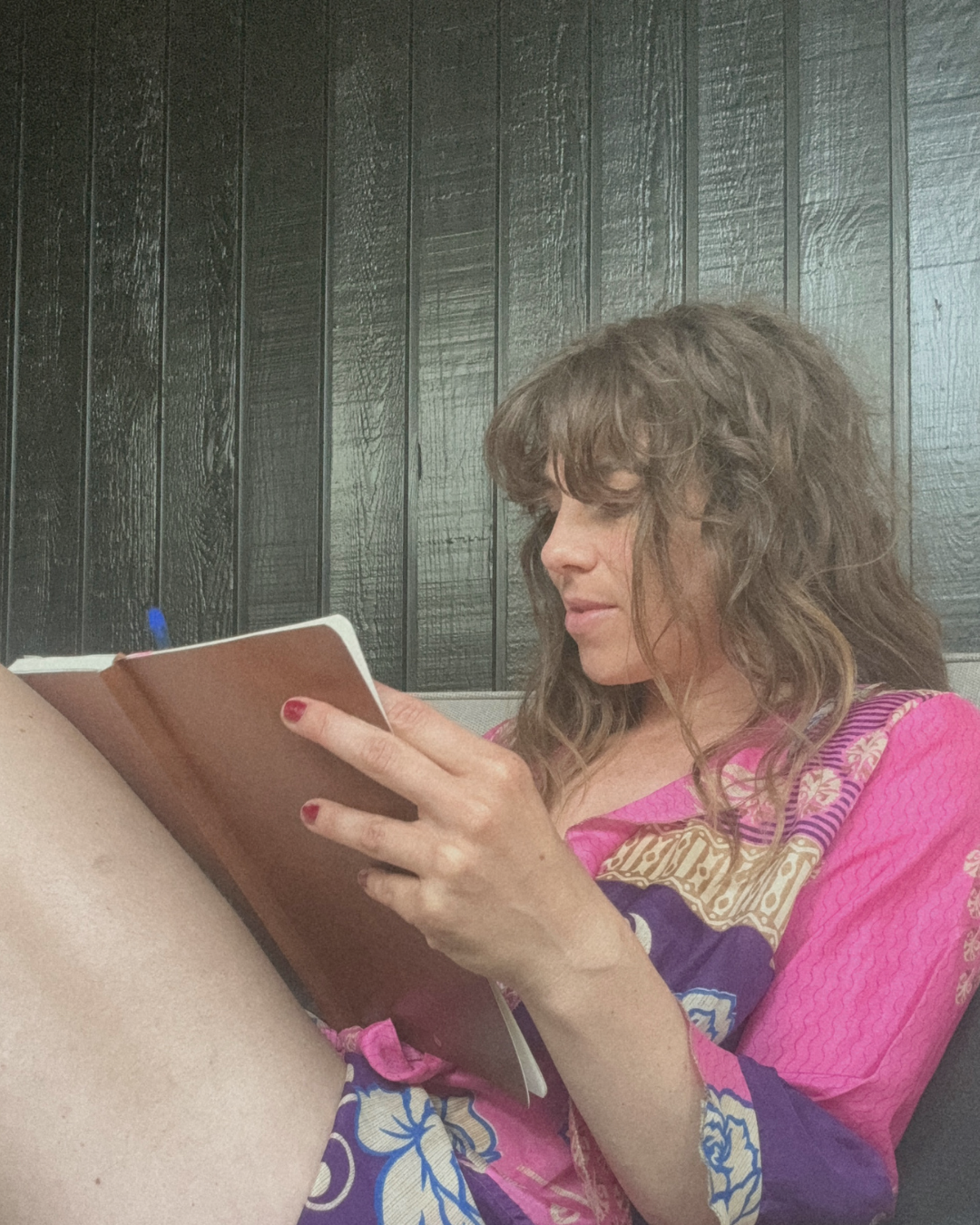 A woman with curly brown hair and pink nail polish is sitting against a dark wooden wall, reading a small notebook while holding her leg.