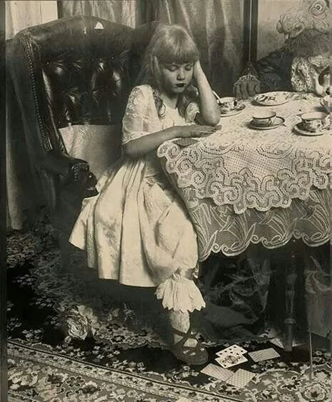 A young girl with long hair and a dress sitting at a table with a lace tablecloth, holding her head with one hand and looking at a book or paper. The table has cups and a teapot, and there is a doll or stuffed animal with a tiger head next to her.