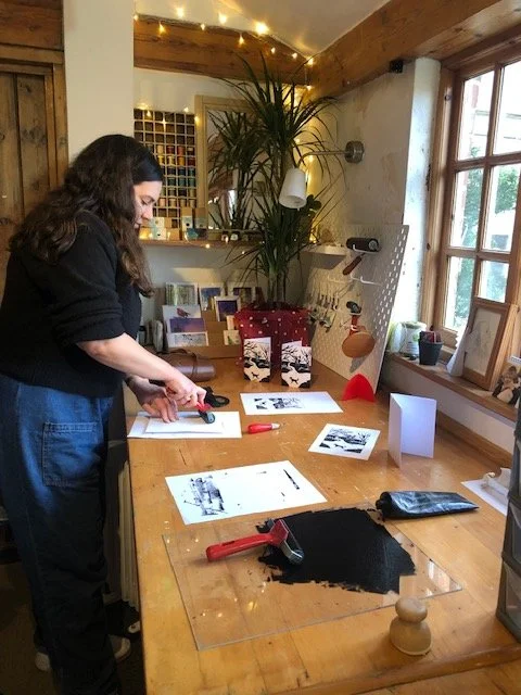 A woman with dark hair learning about lino-cutting in a cozy, well-lit room with a large window, houseplants, and decorative items.
