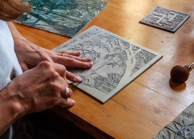 A person is working on a metal embossing or printmaking plate with detailed nature imagery, using a small stamping tool on a wooden table, with other plates and tools nearby.