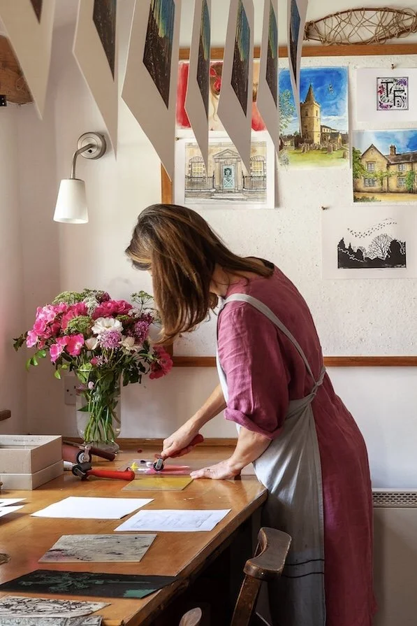 A woman with brown hair wearing a pink dress and apron stands at a wooden table in a room decorated with paintings. She is using a hot glue gun on a small object, with a bouquet of pink and white flowers nearby.