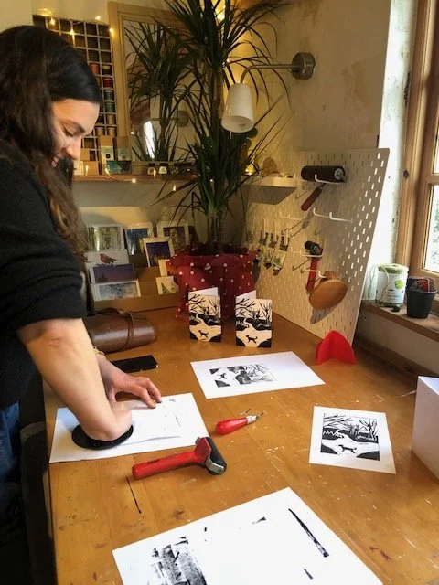 A person working at a wooden table with printing tools and paper, in a cozy room with a window, houseplant, and pegboard with tools and decorations.