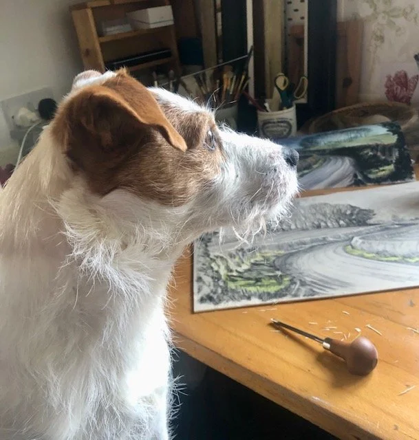 Dog with brown and white fur looking to the right on a wooden table with paintings and art supplies.
