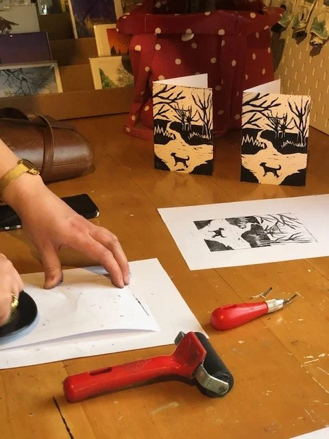 Making linocuts for Christmas cards featuring a black silhouette of a deer and trees, with linocutting and printing tools on a wooden table.