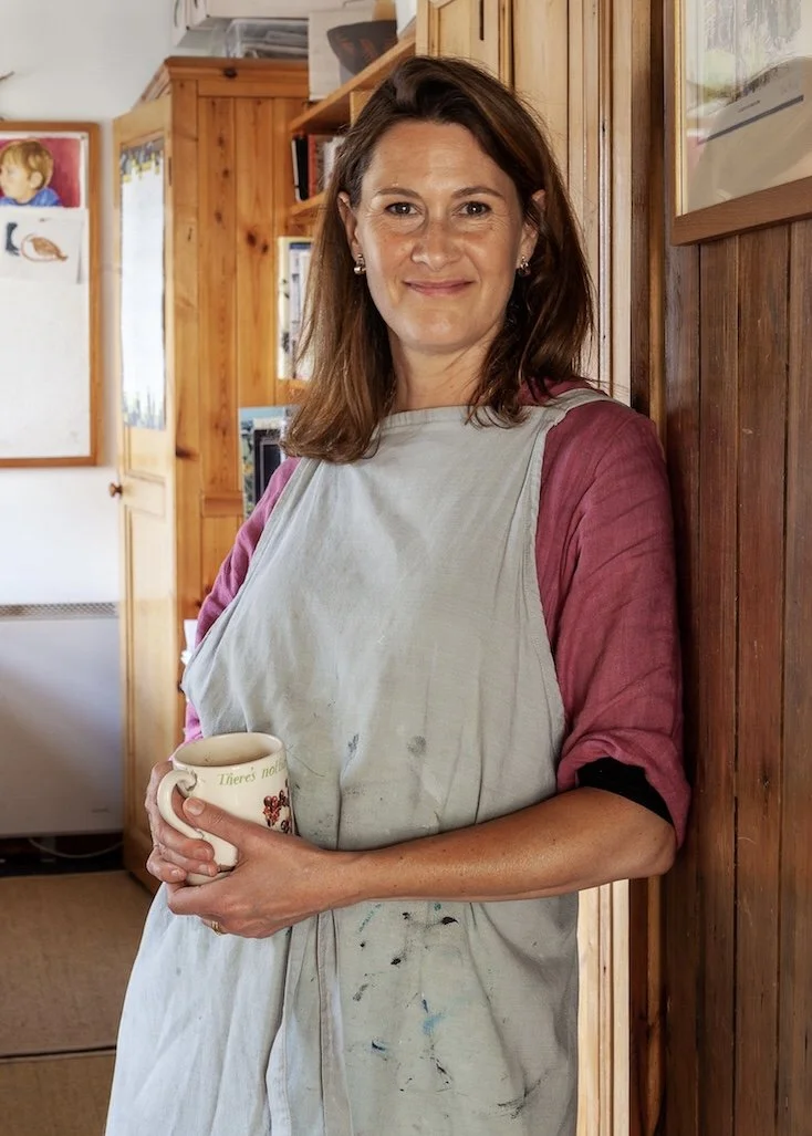 A woman with shoulder-length brown hair wearing a grey apron and a maroon shirt standing indoors, holding a coffee mug, smiling, with wooden cabinets and colorful drawings on the wall in the background.