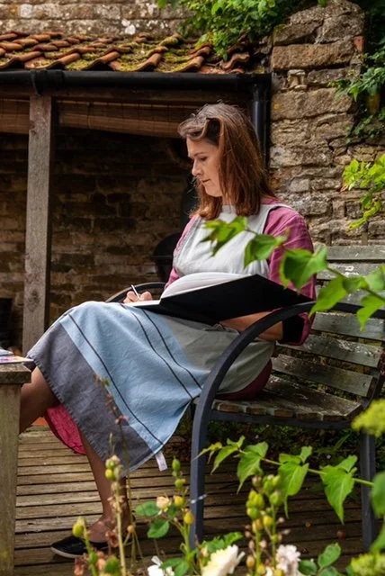 Woman sitting on a garden bench, drawing on a large sketchbook outdoors surrounded by plants and a stone wall.