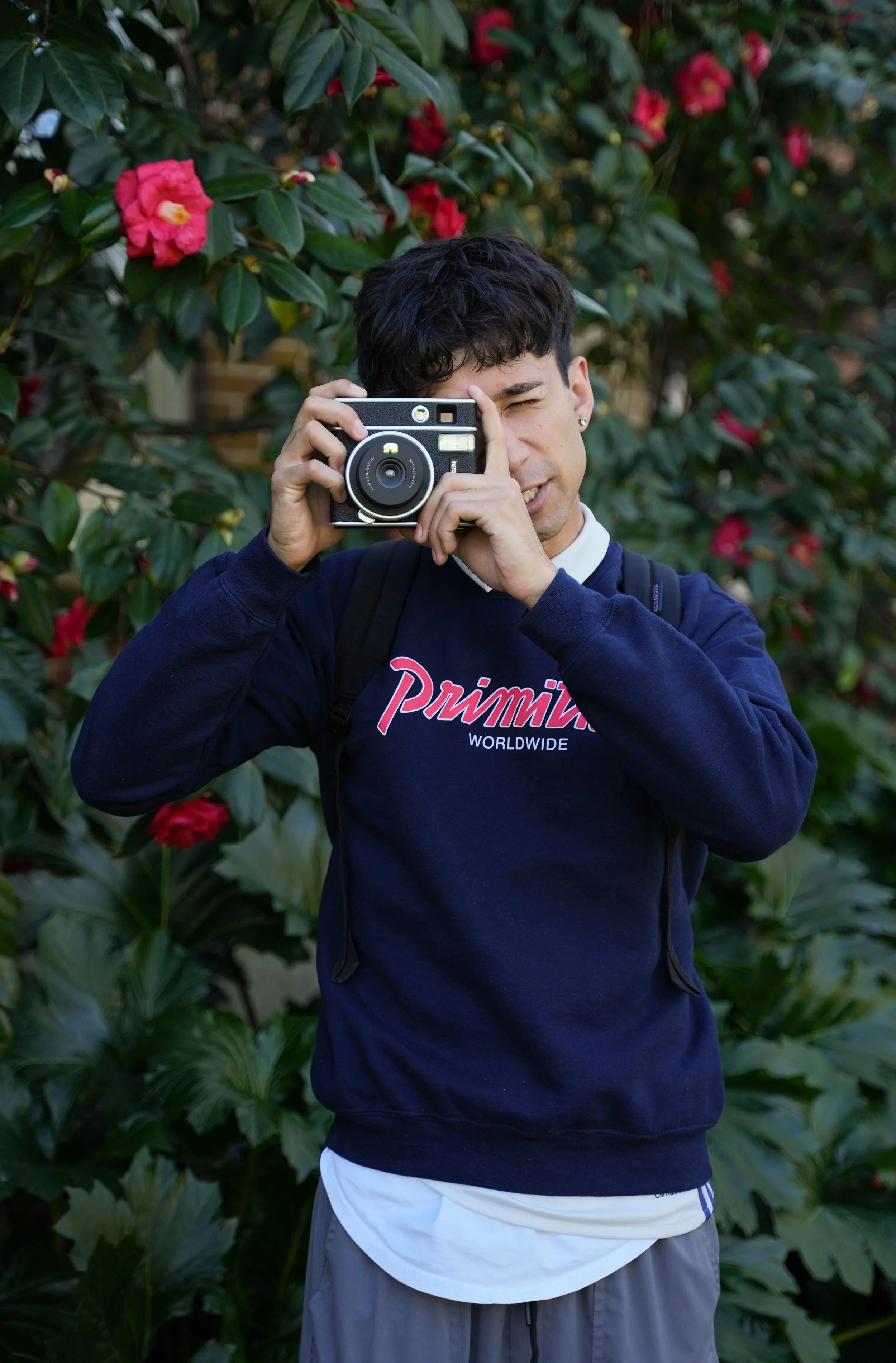 Person taking a photo outdoors with a vintage camera, wearing a navy sweatshirt and standing in front of blooming bushes.