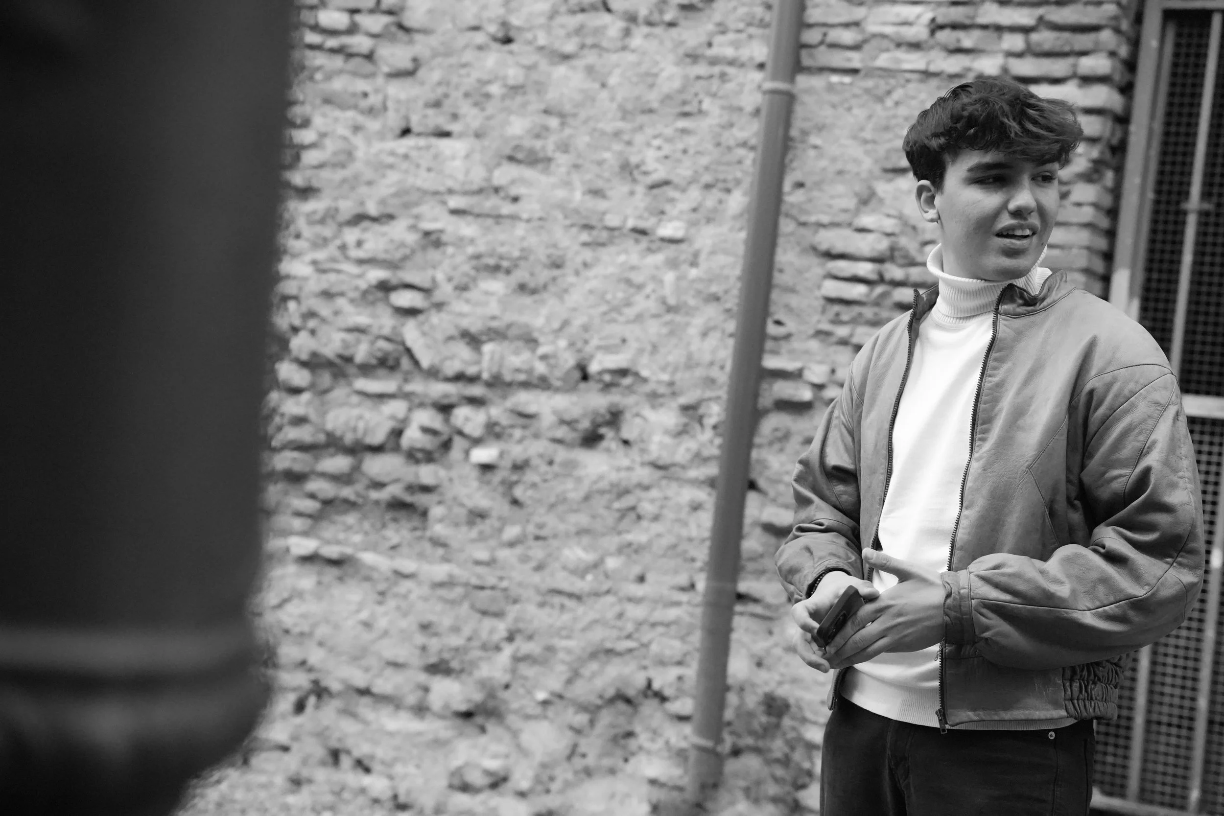 Black and white photo of a person in a jacket standing near a brick wall, holding a smartphone.