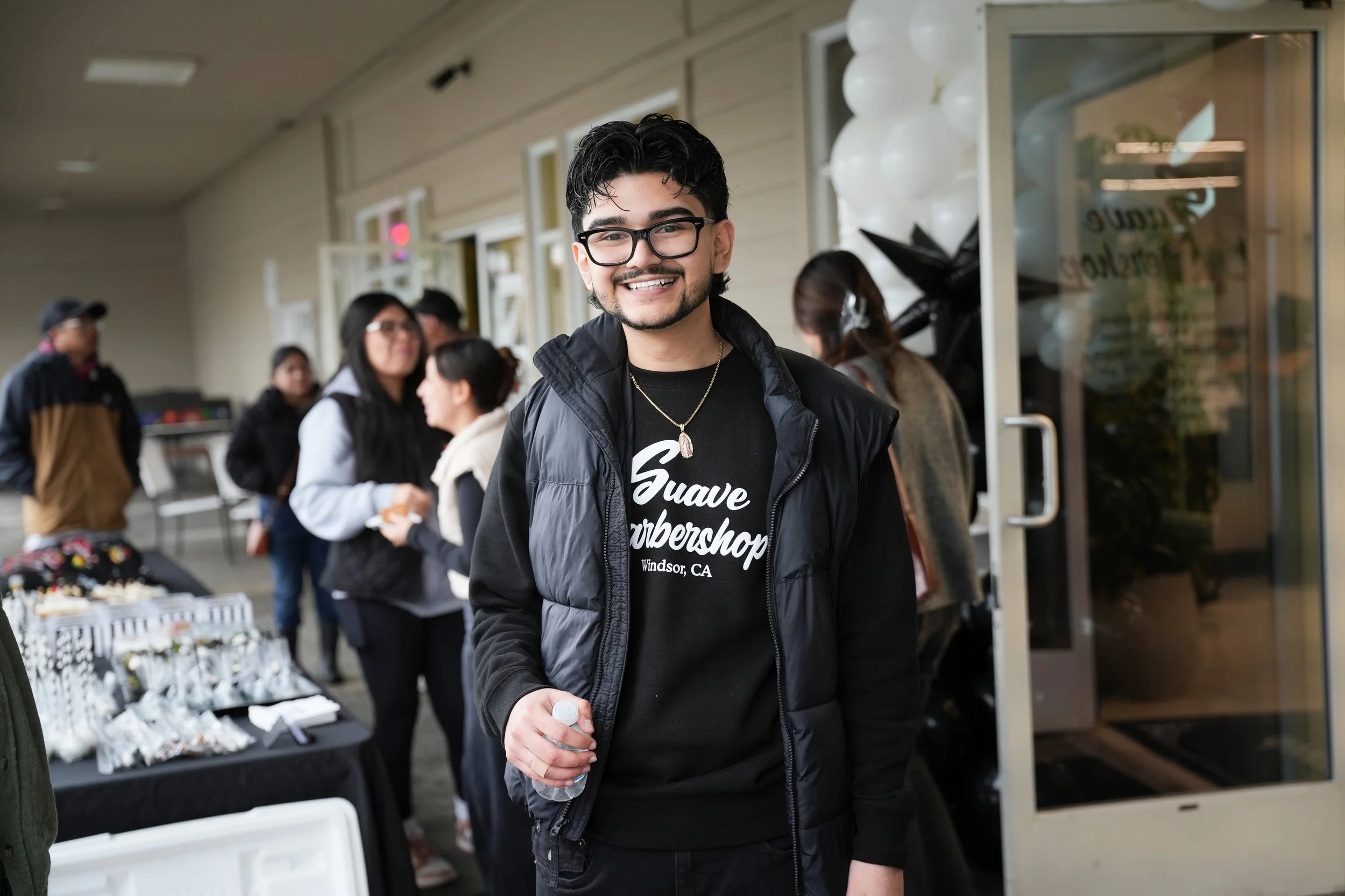 Man wearing glasses and black jacket smiling in front of a storefront, with people and a table with refreshments in the background.