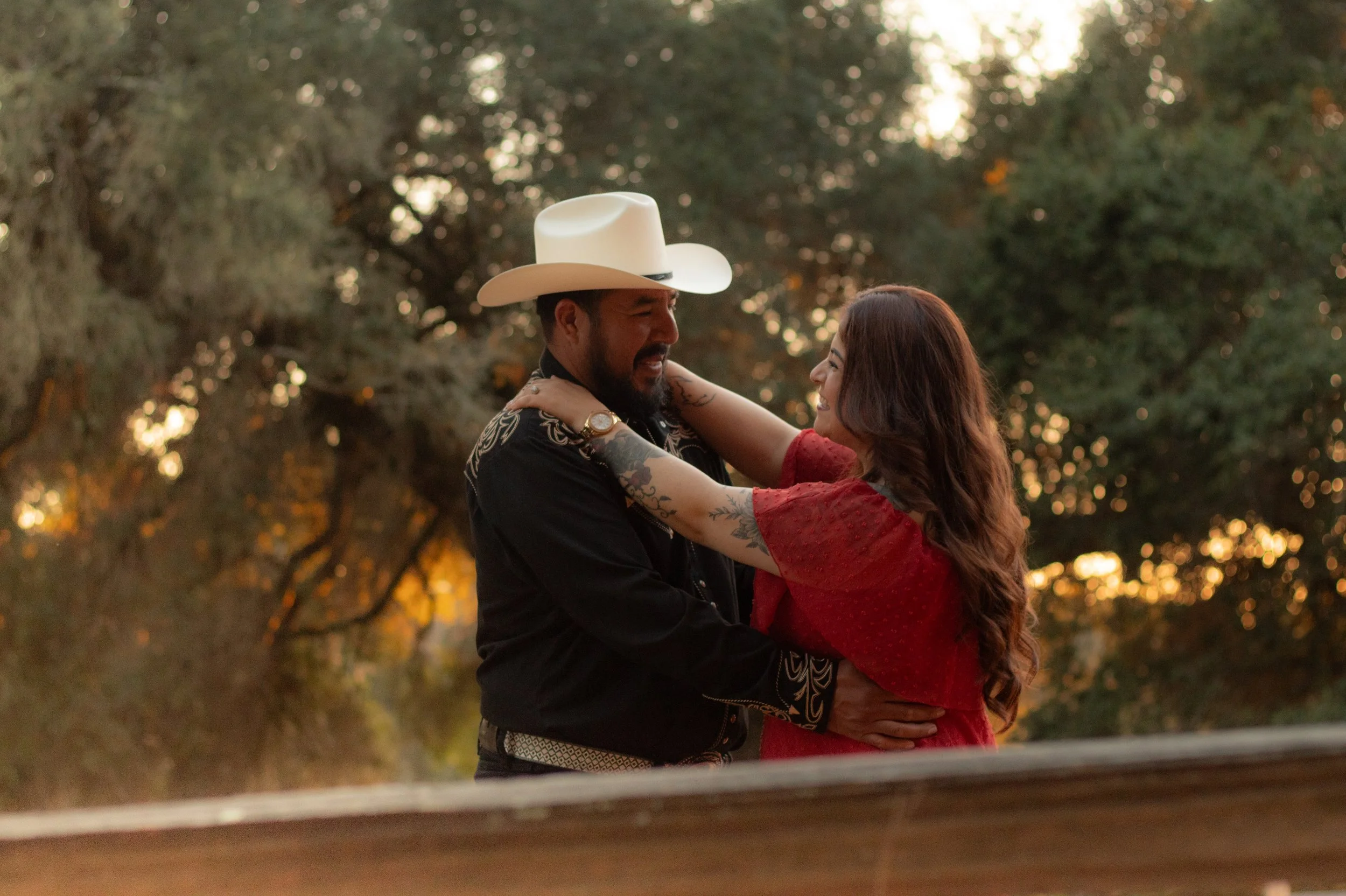 A couple embraces in an outdoor setting. The man wears a white cowboy hat and a black embroidered shirt, while the woman has long hair and wears a red dress. Trees and warm lighting are in the background in Sebastopal California