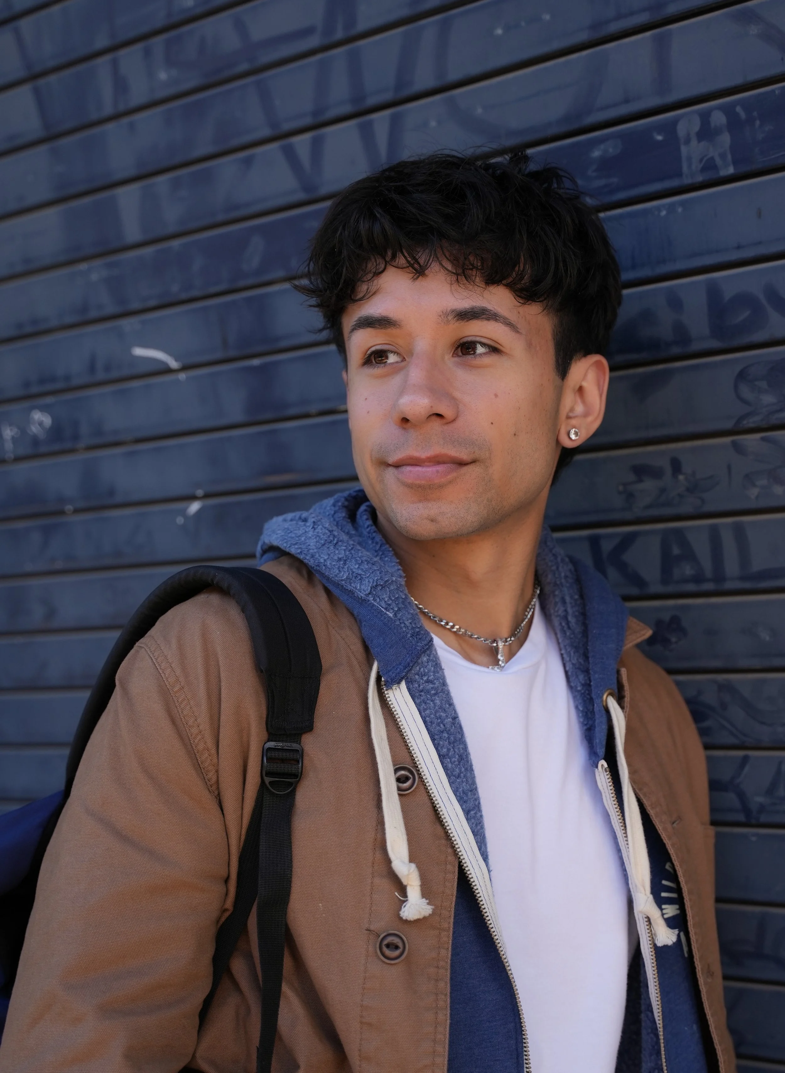 Young man with short dark hair wearing a brown jacket, blue hoodie, and white t-shirt, standing against a blue graffiti-covered wall, looking to the side with a slight smile at Santa Rosa Junior College.