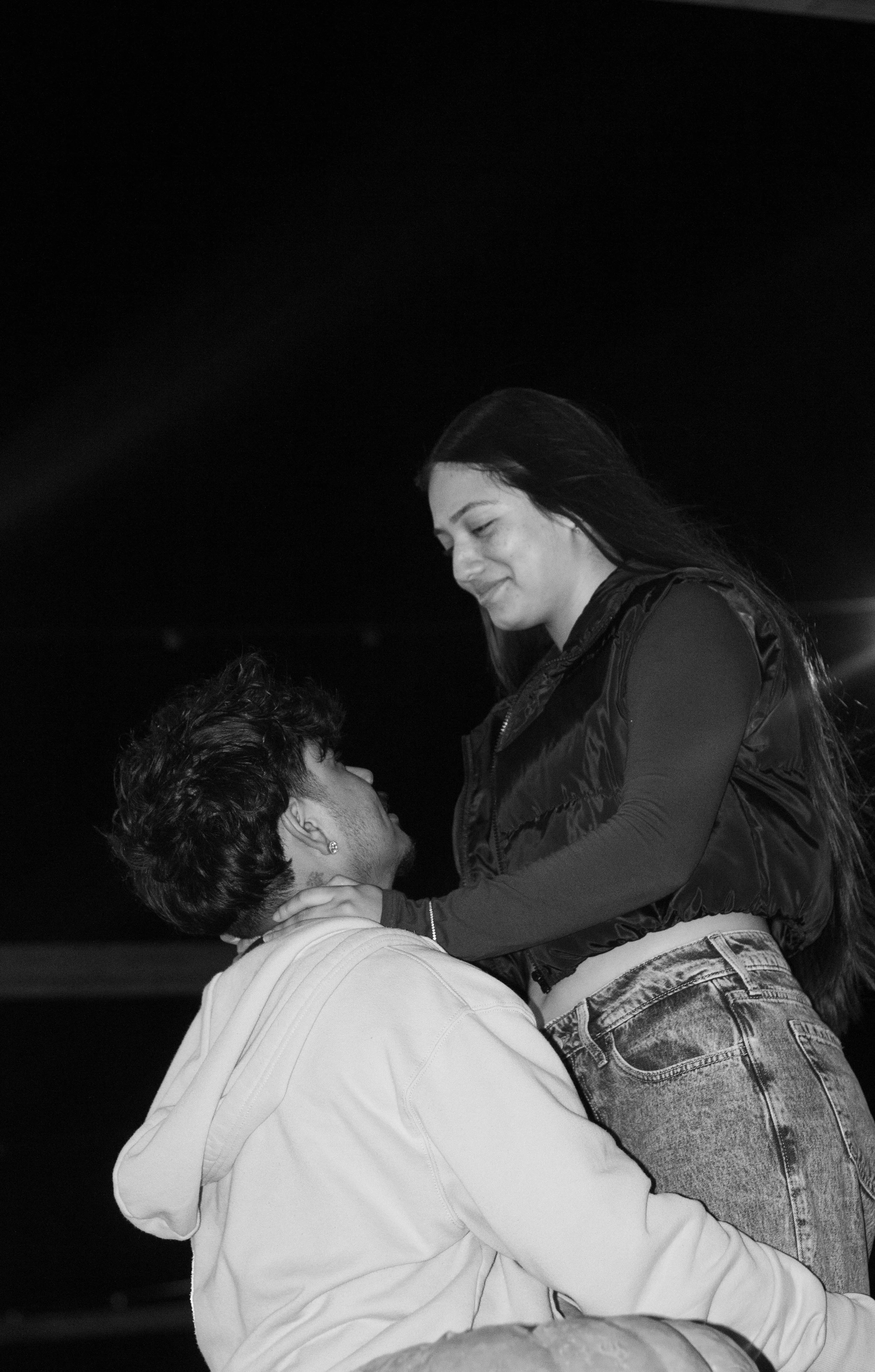 A black and white photo of a couple embracing. The woman is standing and smiling down at a kneeling man holding her close.