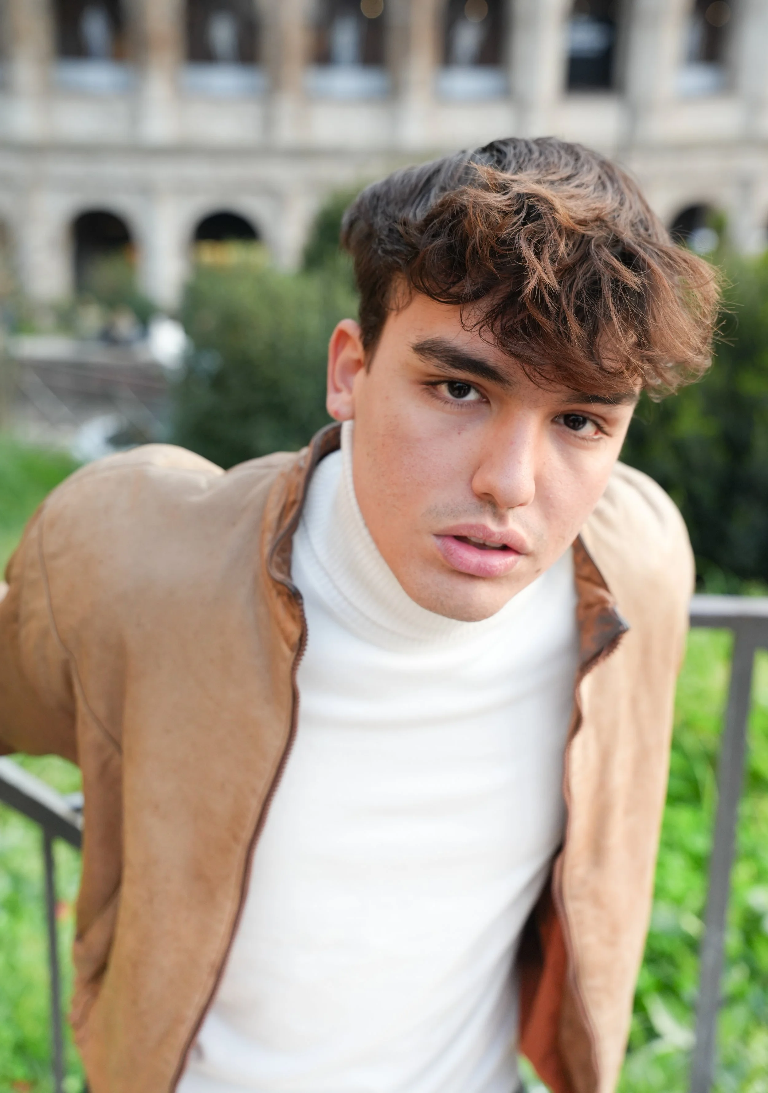 Young man wearing a beige jacket and white turtleneck, with the Colloseum in the background and greenery.