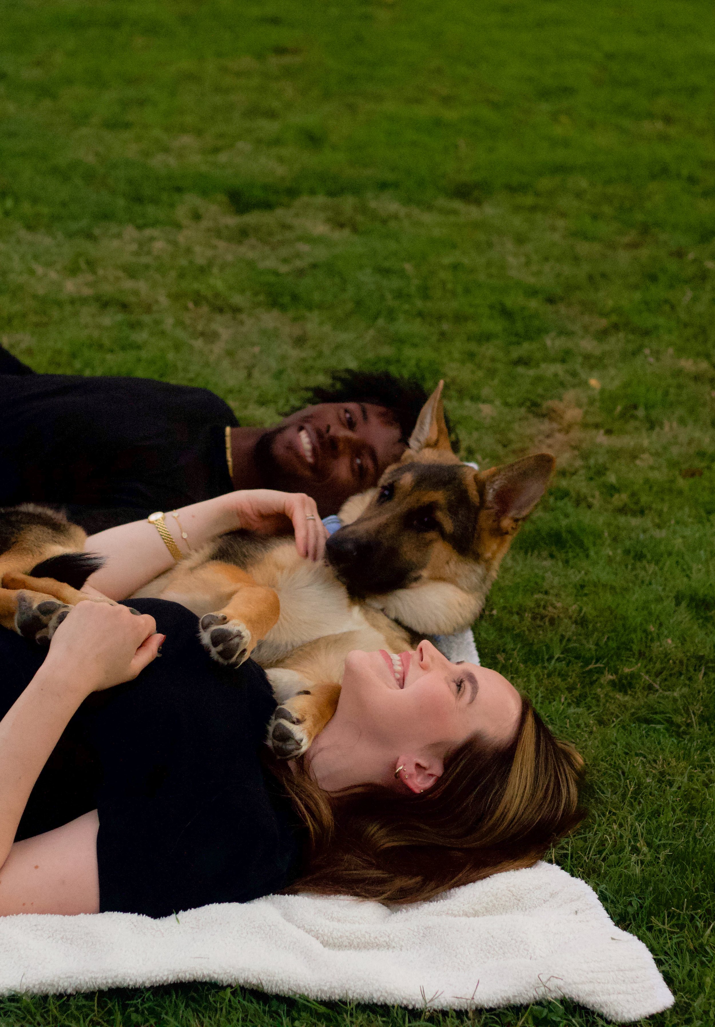 Two people lying on the grass with a German Shepherd dog, all appearing relaxed and happy.