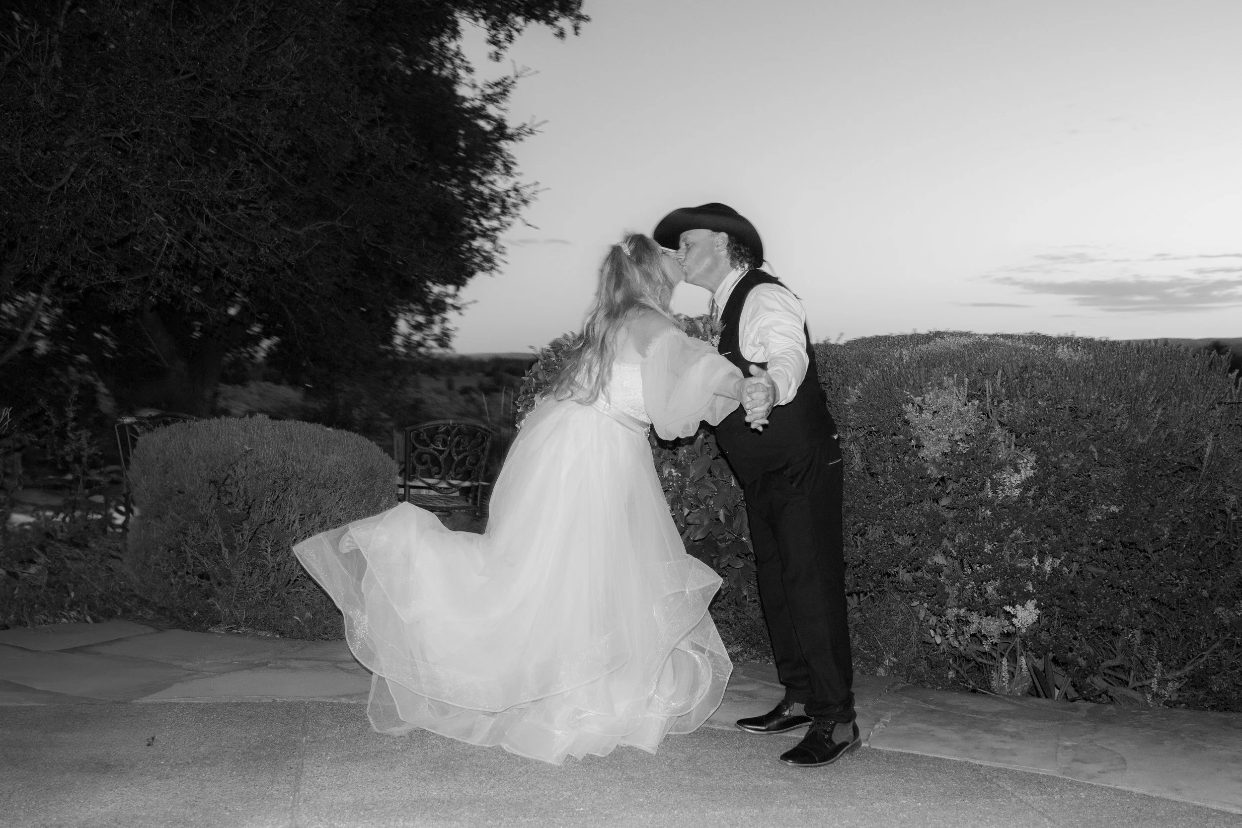 A couple dressed in wedding attire kissing outdoors, with the bride in a flowing gown and the groom in a cowboy hat and vest. The setting is a garden with foliage and a sunset in the background.