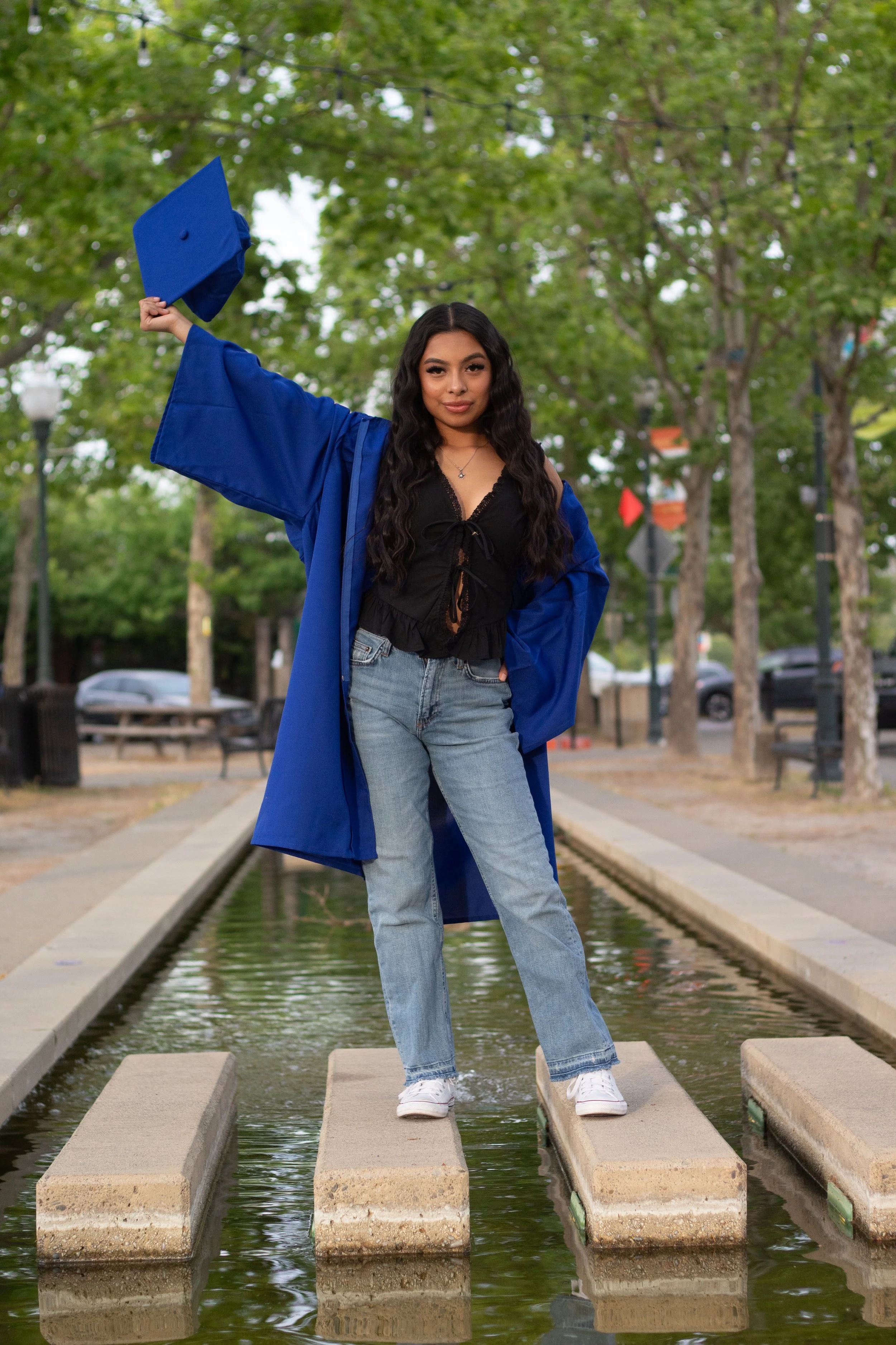 Person in a blue graduation gown and cap standing on concrete stepping stones in a shallow water feature, surrounded by trees.
