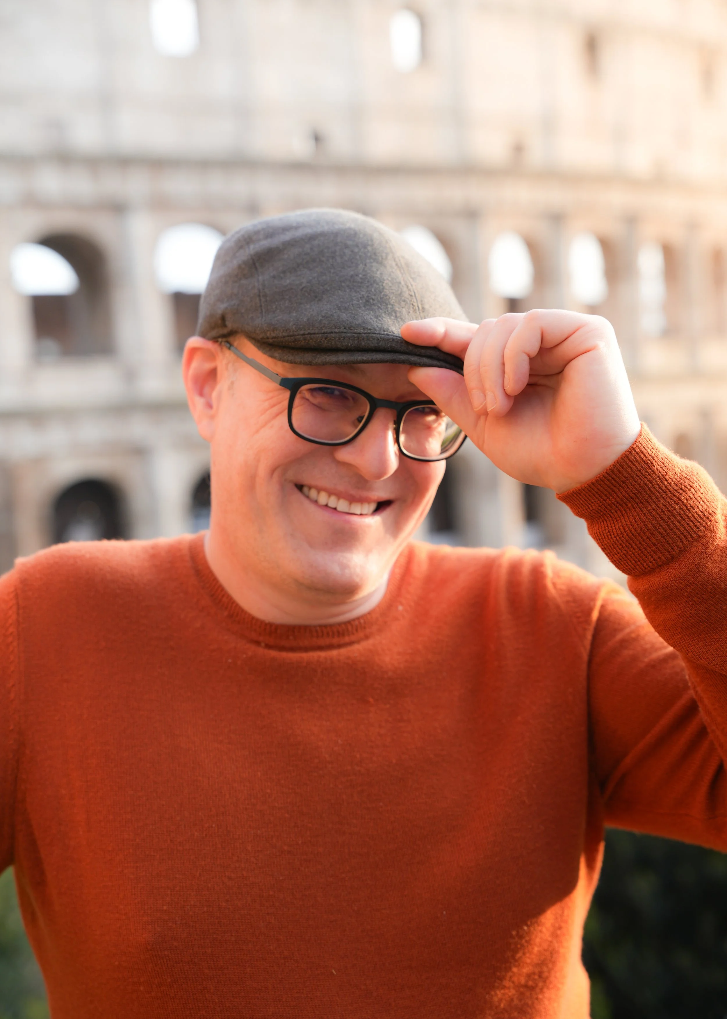 Man in orange sweater and flat cap smiling at the Colosseum.