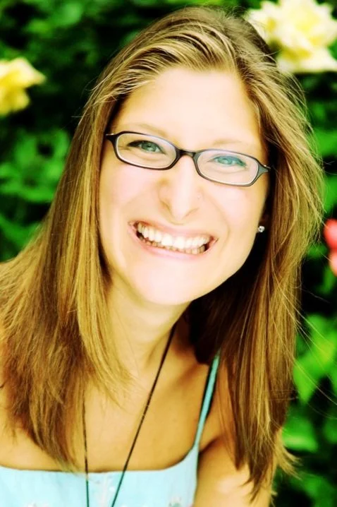 Smiling woman wearing glasses with long hair, against a blurred green and floral background.
