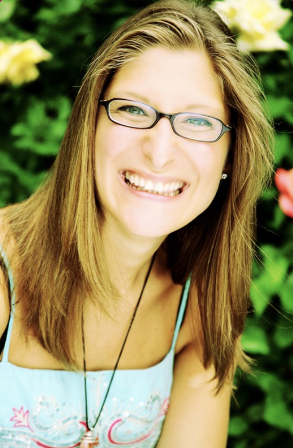 Person with long hair and glasses smiling against a background of greenery and flowers.