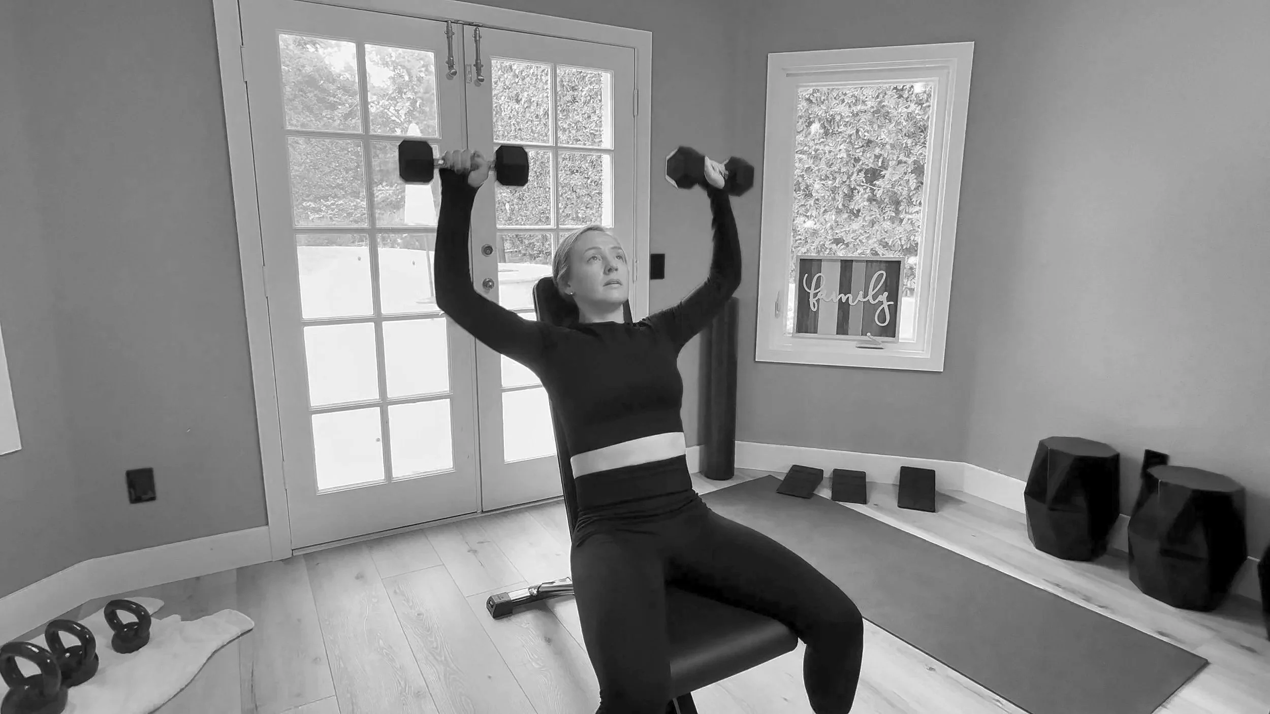 A woman in workout attire is seated on a chair, lifting dumbbells with her arms raised. The room has a yoga mat, kettlebells, and other fitness equipment on the floor, with a window and a door behind her, and a sign that says 'family' on the wall.