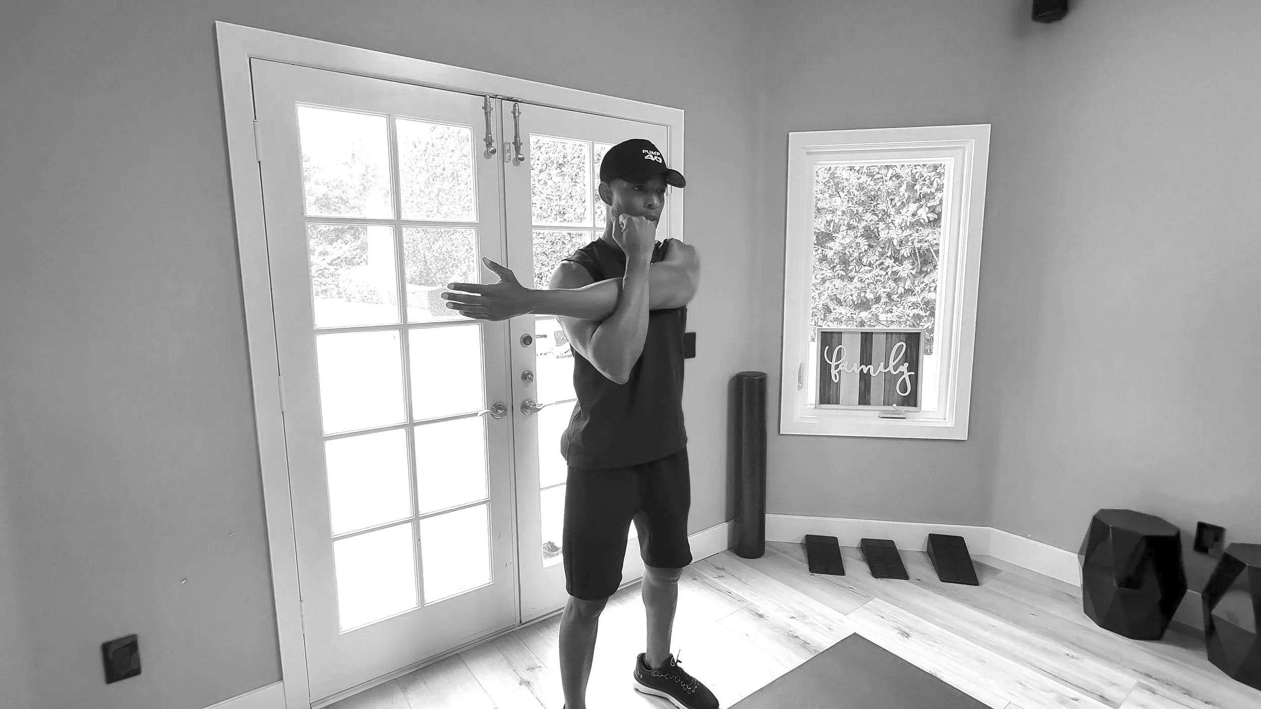 A person in workout attire stretching inside a room with large windows, a door, and exercise equipment. The person is holding one arm across their chest and has their other hand near their chin.