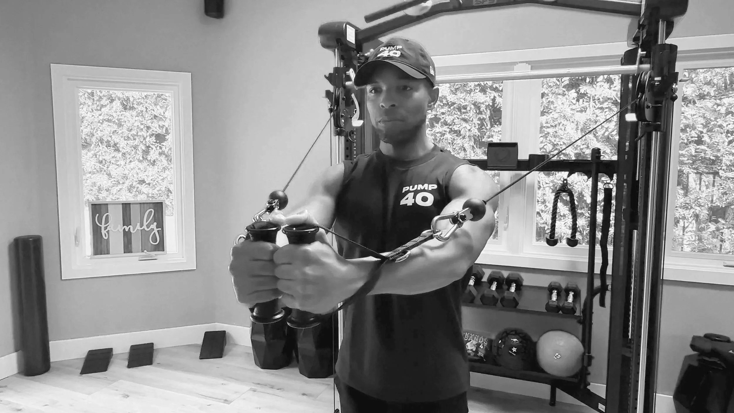 Man wearing a cap and sleeveless shirt exercising with resistance cables in home gym, with windows, dumbbells, and fitness equipment in background.