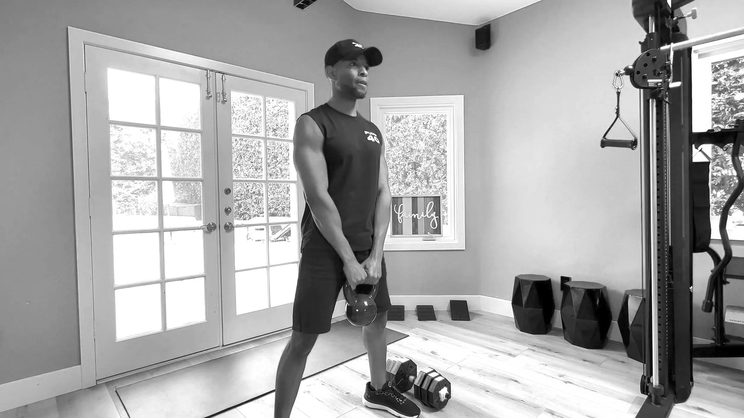 A man in athletic attire performing a kettlebell exercise in a home gym with door and window, fitness equipment, and a framed sign that says 'family' in the background.