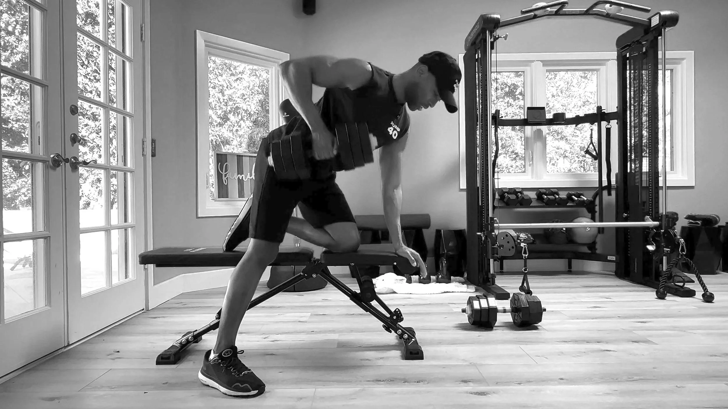 Man performing a bent-over dumbbell row exercise in a home gym with exercise equipment and windows in the background.
