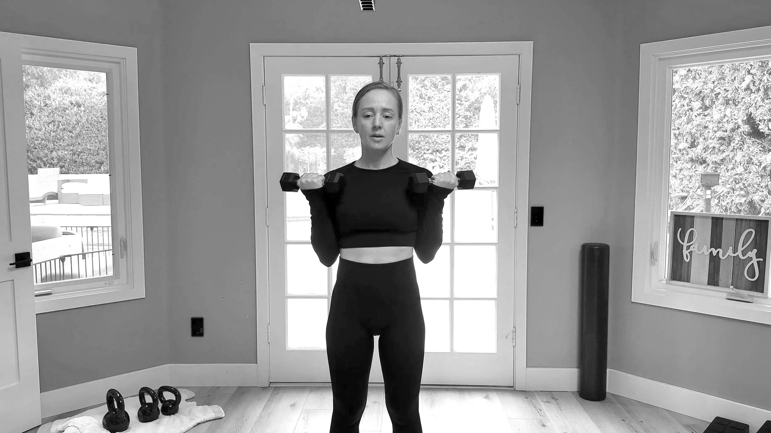A woman in workout clothes holding dumbbells in a home gym with windows and exercise equipment.