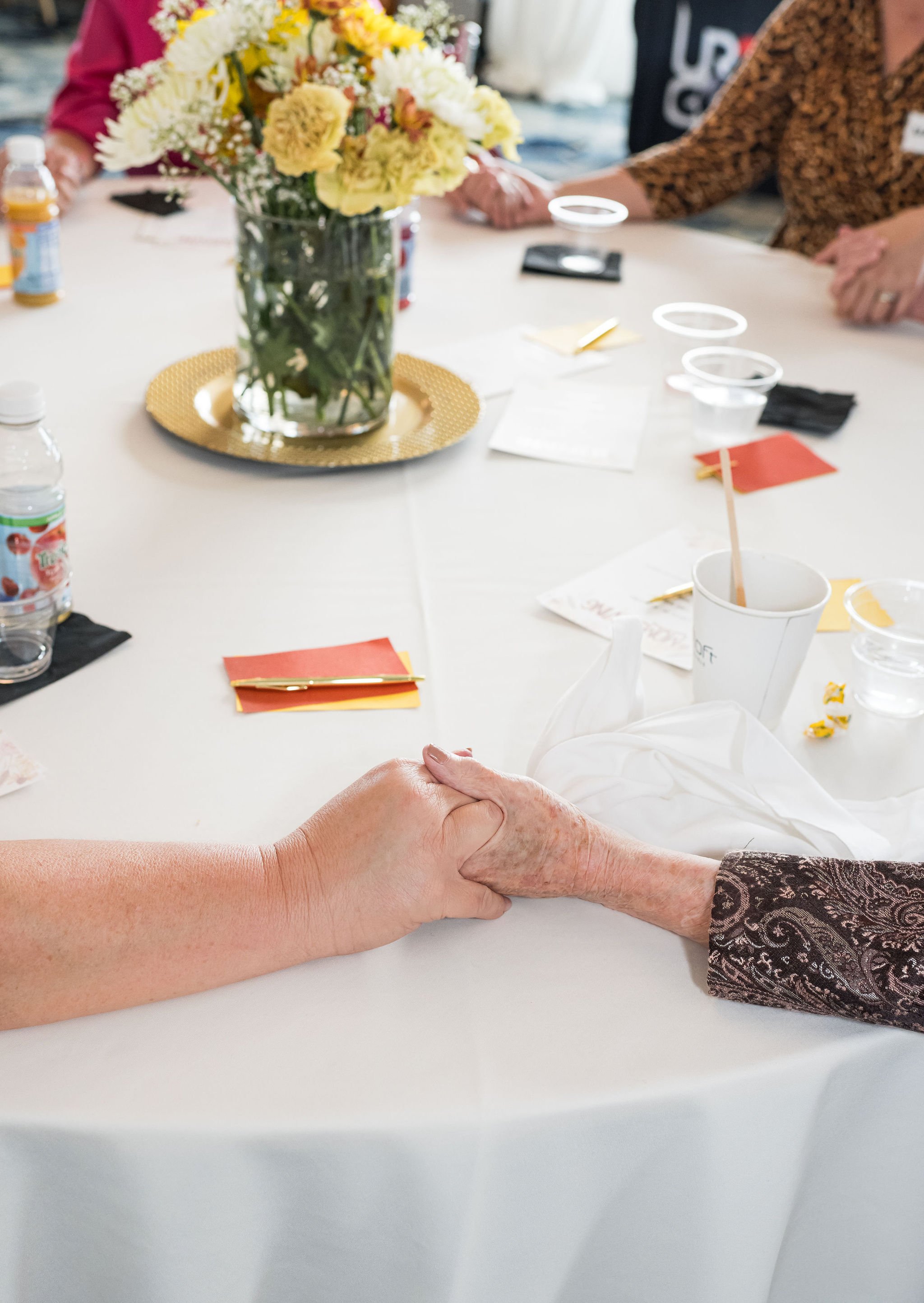 Hands holding across a table, with a vase of flowers and personal items scattered around.