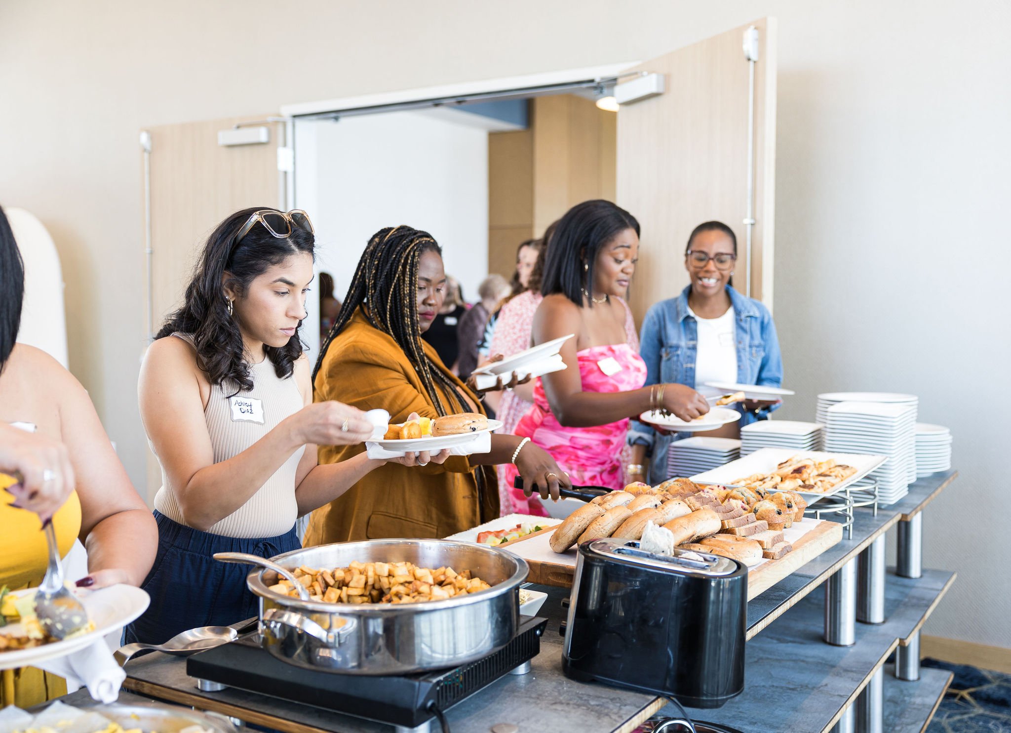 People serving themselves food at a buffet with dishes like bread, potato cubes, and pastries.