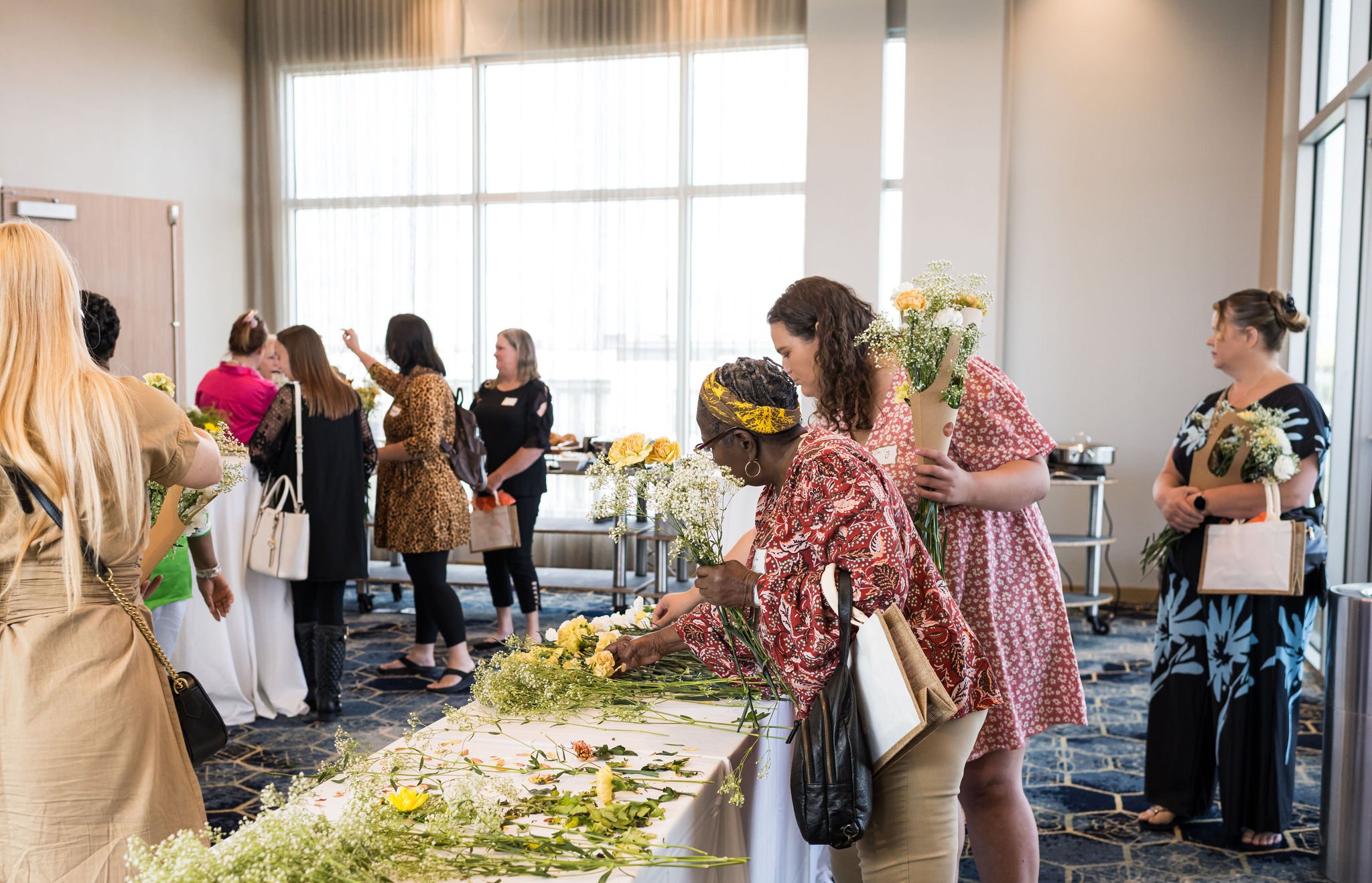Women arranging flowers in a room with large windows, some holding bouquets and others picking flowers from a table.