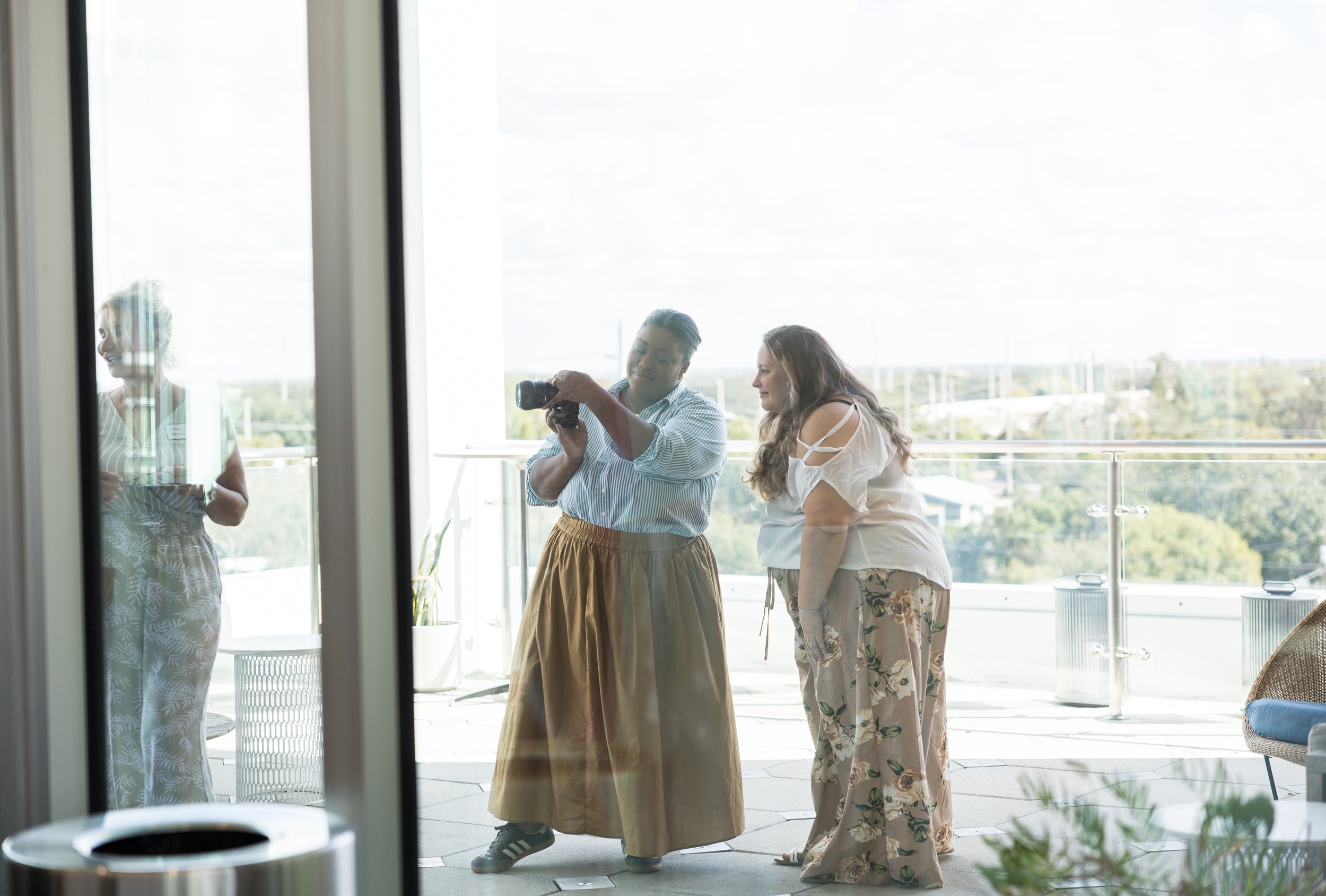 Two people on a balcony reviewing photos on a camera, with a reflection of a third person holding a plate seen through glass. The balcony has clear glass railings and overlooks greenery.