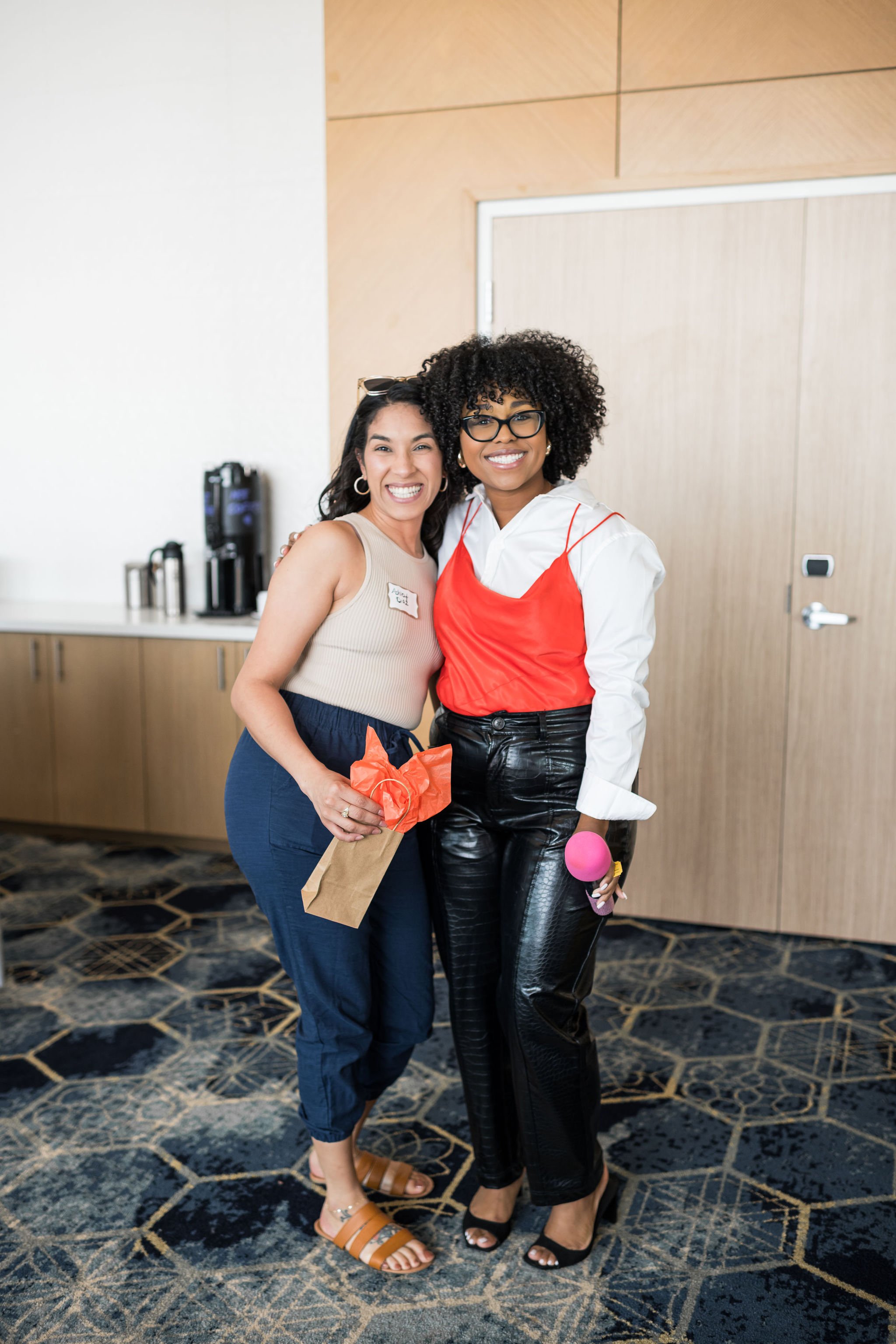Two women smiling and posing together indoors, one holding a paper bag and the other a pink microphone, with a coffee station in the background.