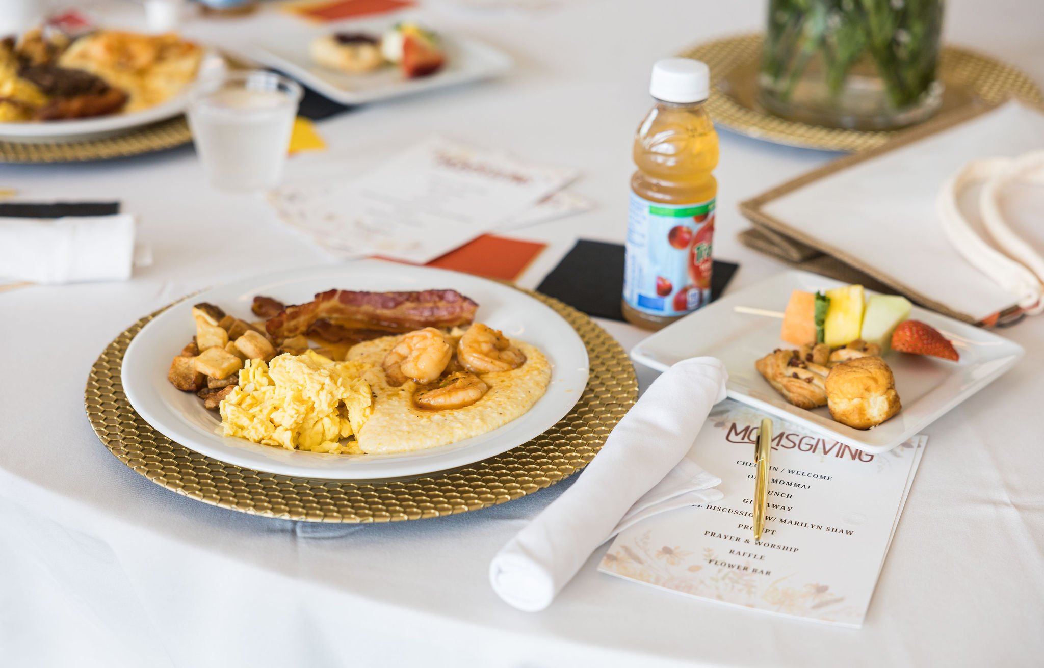 Brunch table setting with scrambled eggs, bacon, shrimp, grits, roasted potatoes on a white plate, fruit skewer and pastries on a side plate, apple juice bottle, and a MomsGiving event program.
