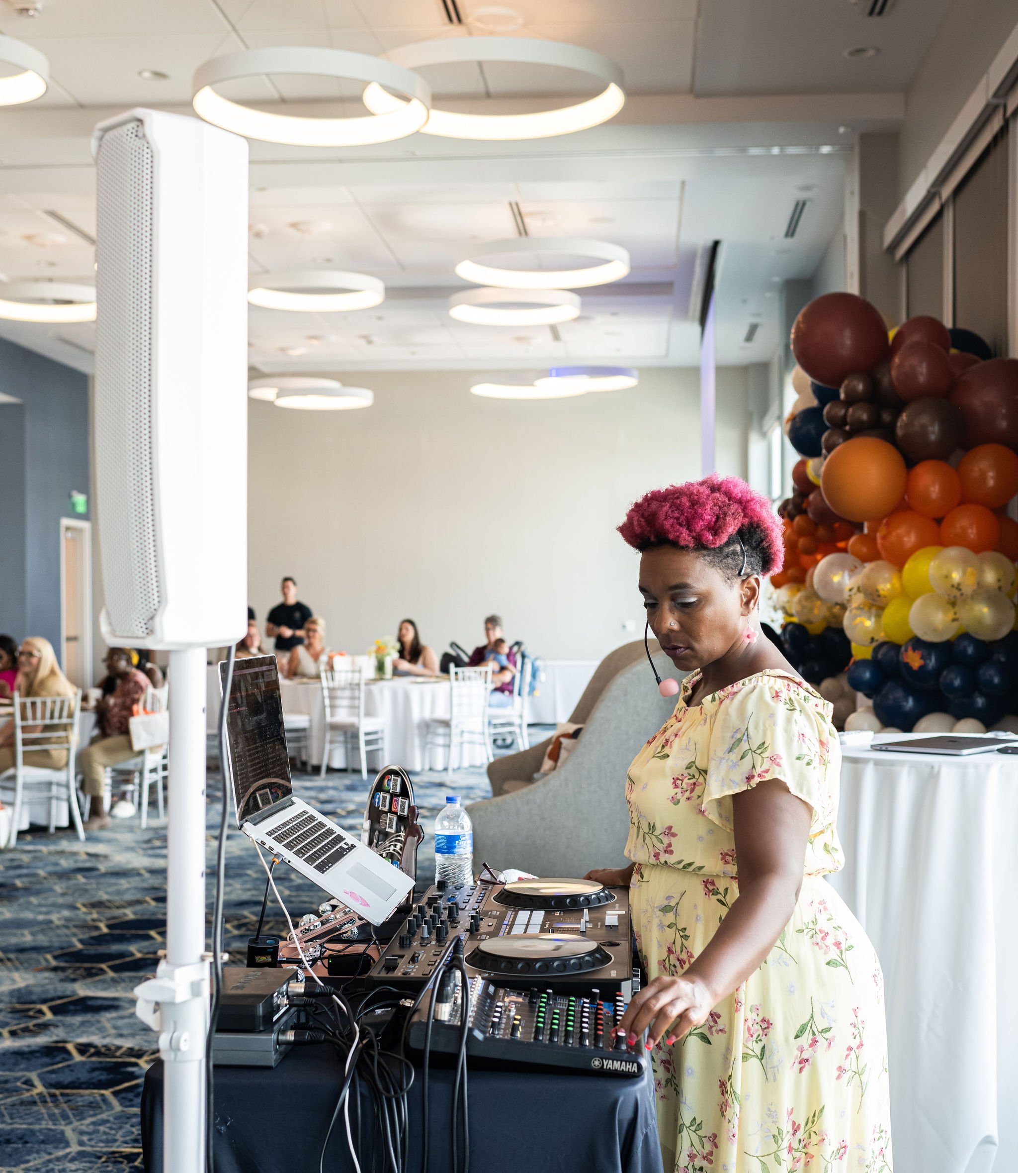 A DJ with pink short hair in a floral dress operates a mixing console at an event. Attendees are seated at tables in the background, and colorful balloons are arranged in the corner. The room has modern lighting and decor.