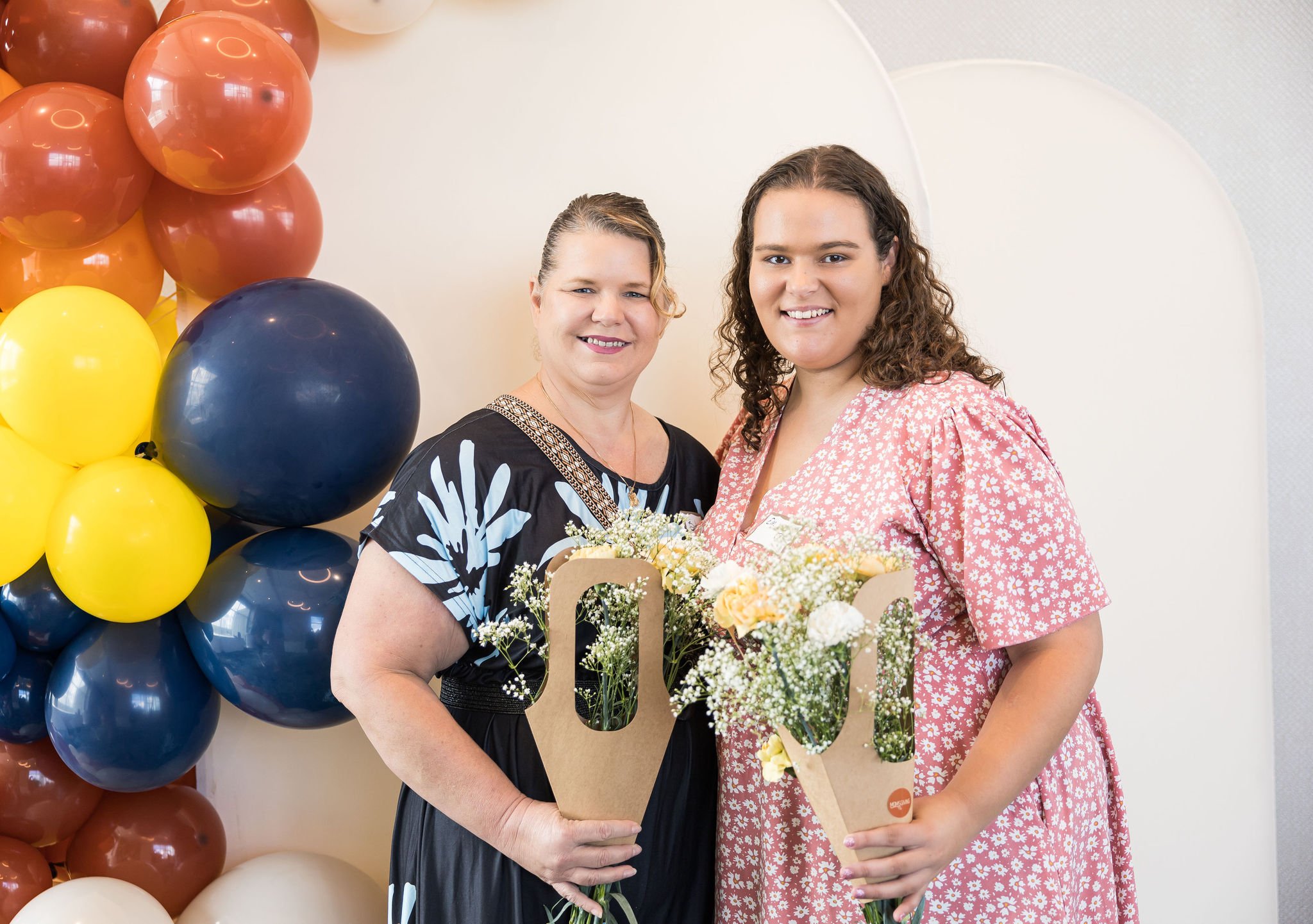 Two women holding bouquets stand in front of a colorful balloon arch.