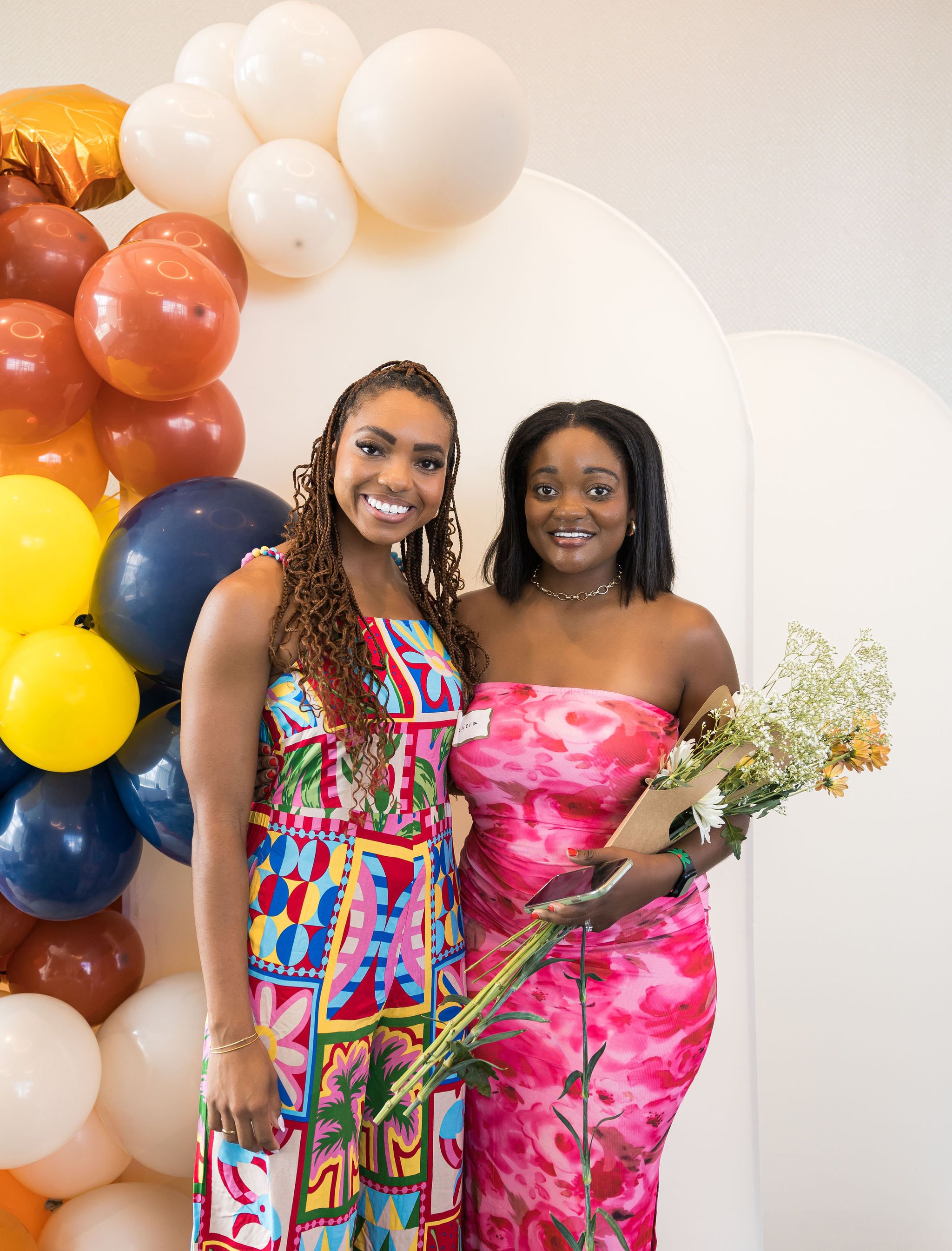 Two women smiling in colorful dresses; one holding flowers, standing in front of a decorative balloon arch.