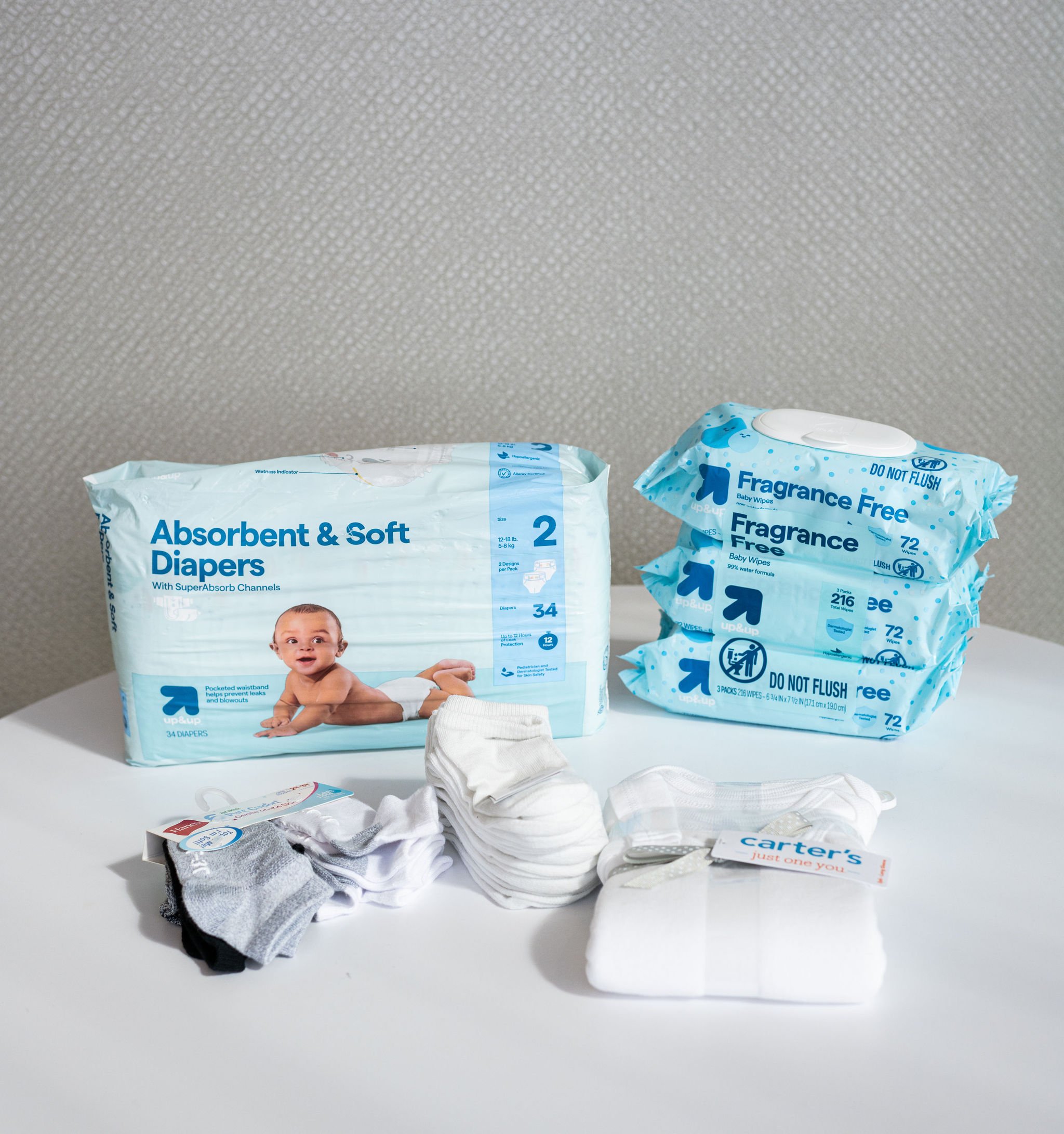 A pack of absorbent and soft diapers, three packs of fragrance-free baby wipes, and a bundle of white baby socks on a table.