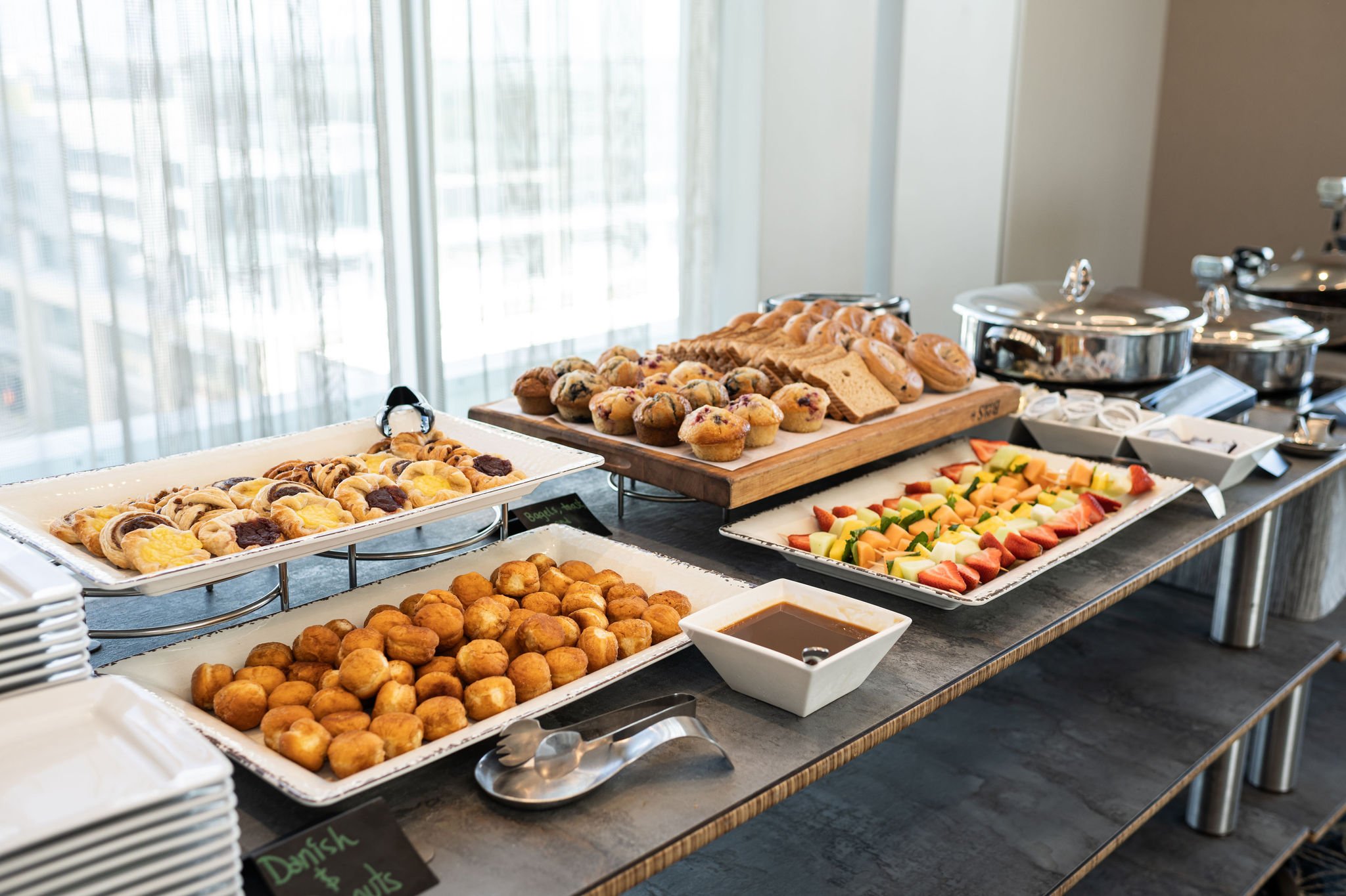 Breakfast buffet with pastries, muffins, fruit skewers, bagels, and sauces in a hotel setting.