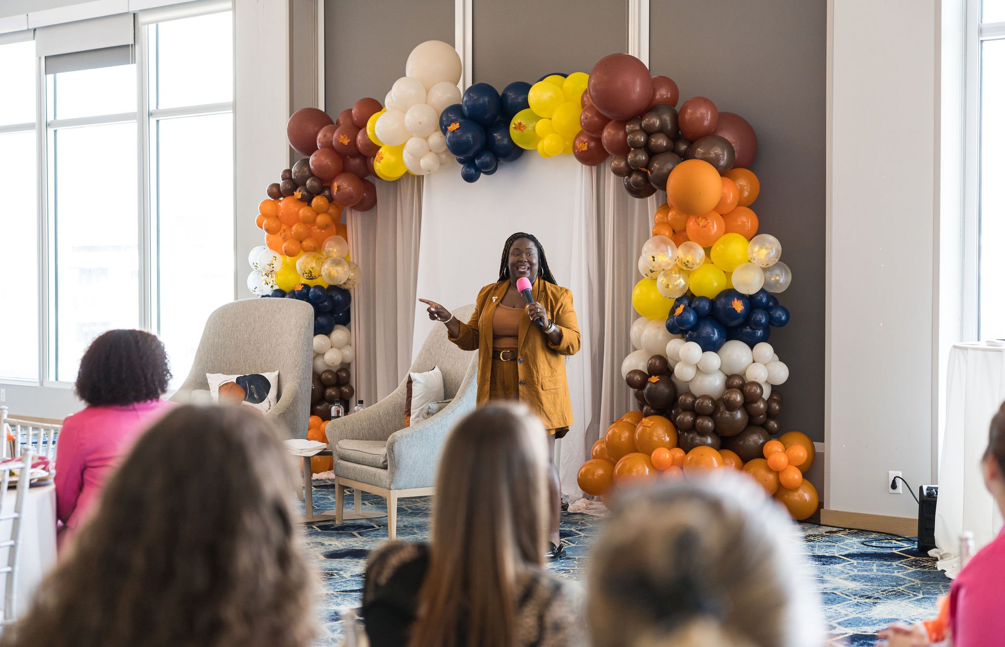 A woman in a brown outfit speaking at an event, holding a pink microphone. She stands in front of a colorful balloon arch with brown, orange, yellow, blue, and white balloons. Audience members are seated in the foreground inside a room with large win