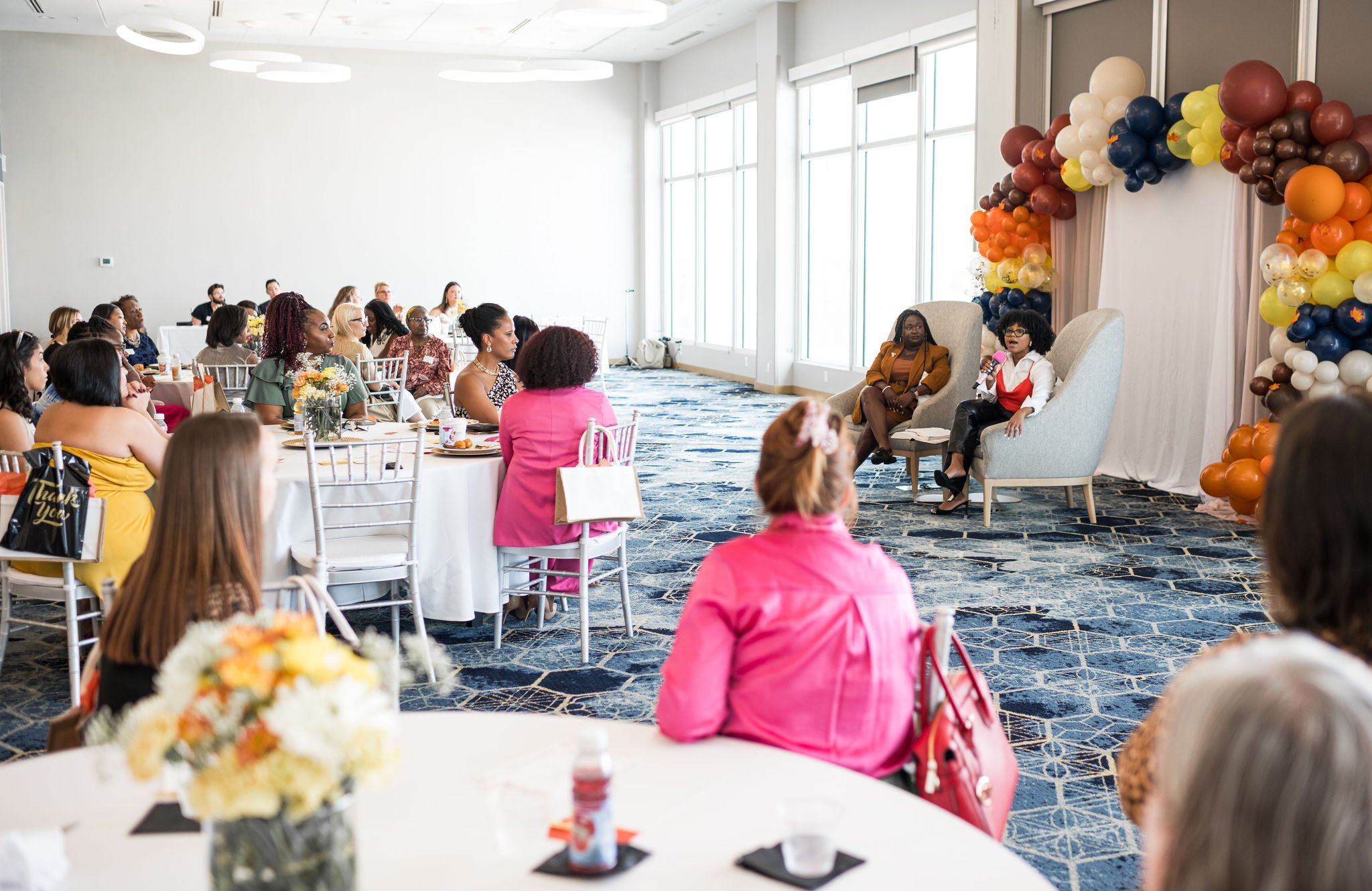 Women attending a conference in a decorated room with balloon arch, speaker on stage, attendees seated at round tables.