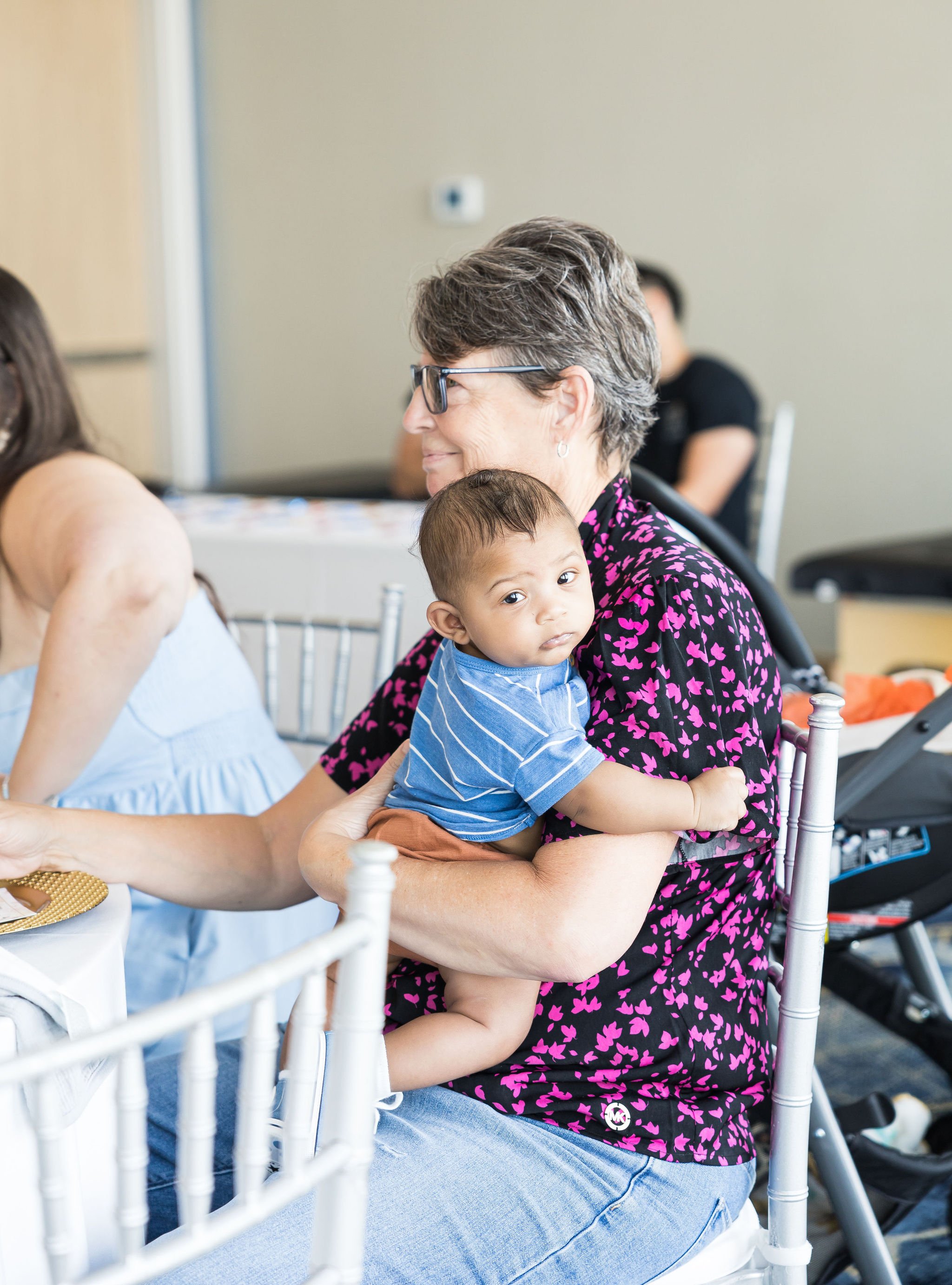 Woman with glasses holding a baby in her arms at an indoor event.