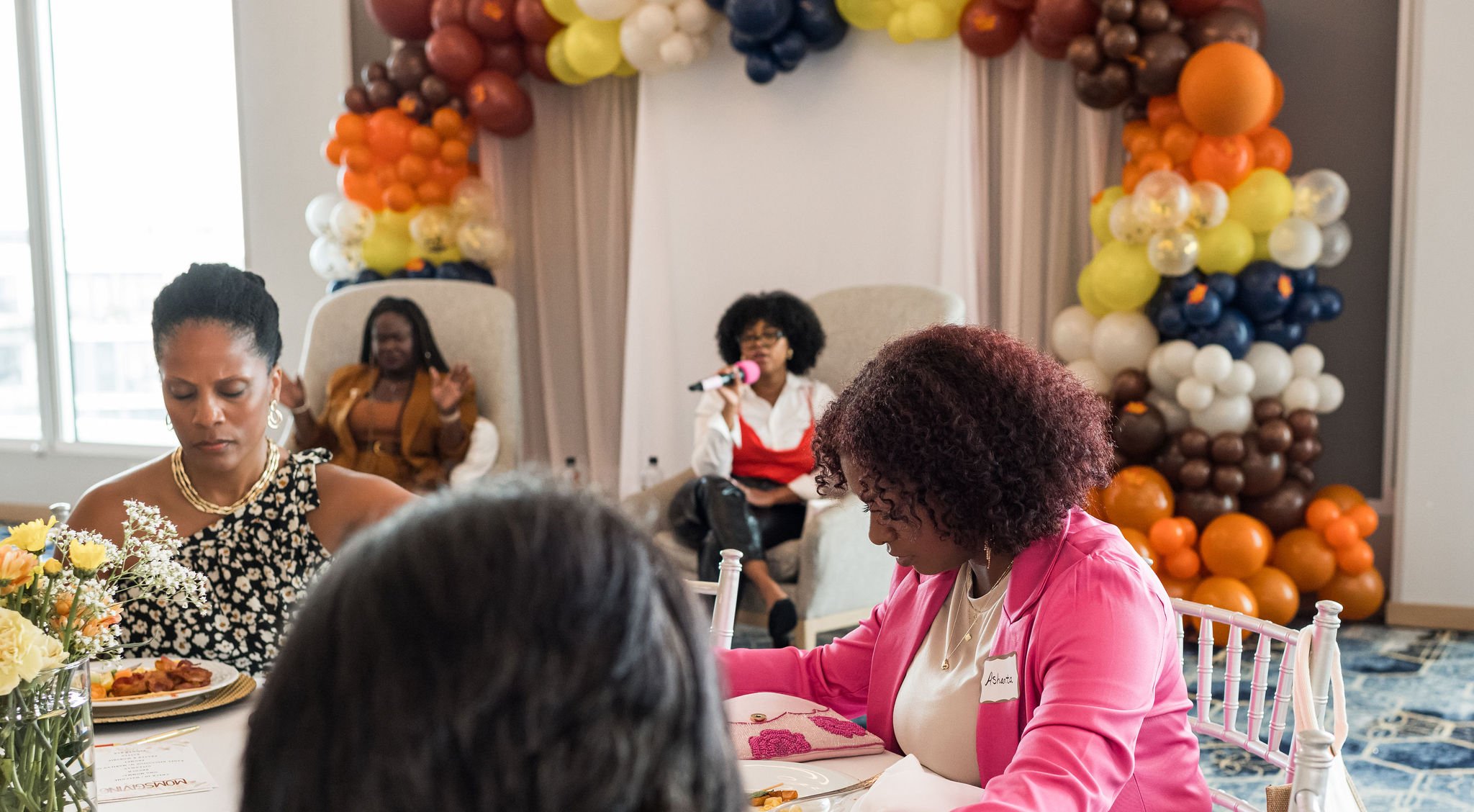 A group of people is seated at a decorated table with flowers and plates. A woman is speaking into a microphone in the background, and there are colorful balloon decorations behind her. The setting appears to be a formal event or gathering.