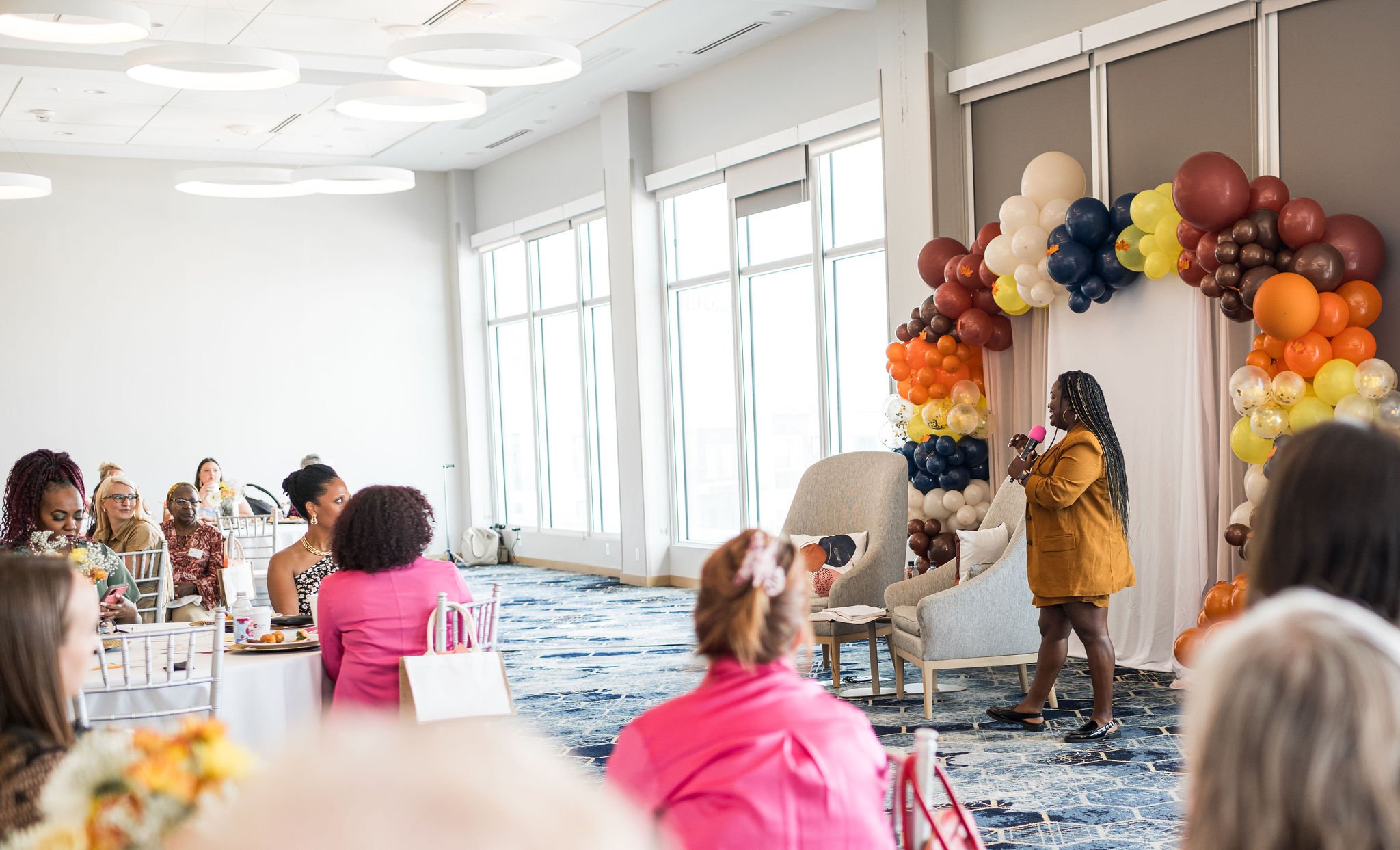 A woman speaks into a microphone at an indoor event decorated with colorful balloons. The audience, seated at round tables with white tablecloths, is listening attentively. The room has large windows and modern lighting.