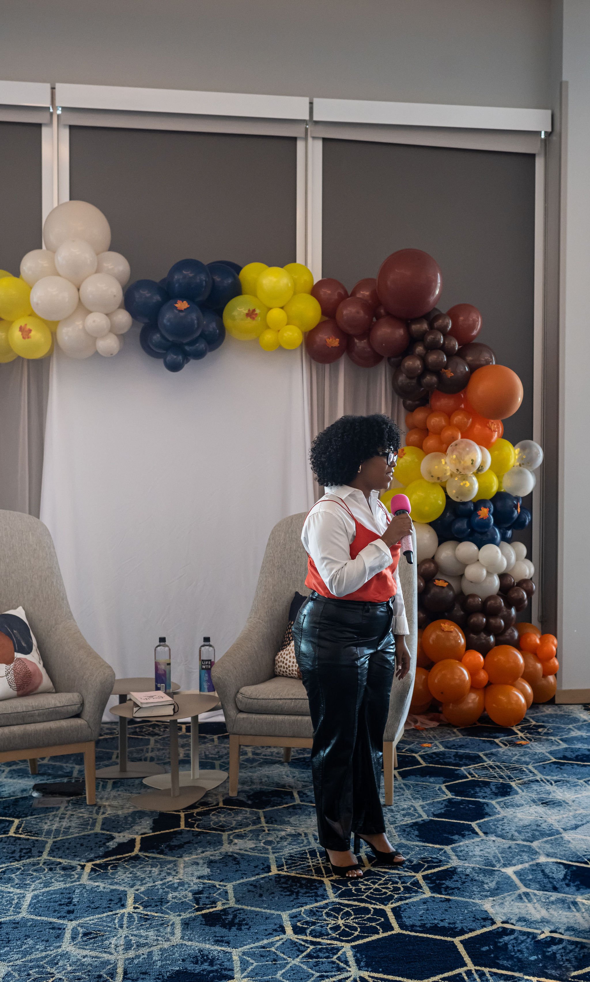 Woman holding a microphone standing in front of a balloon arch at an event.