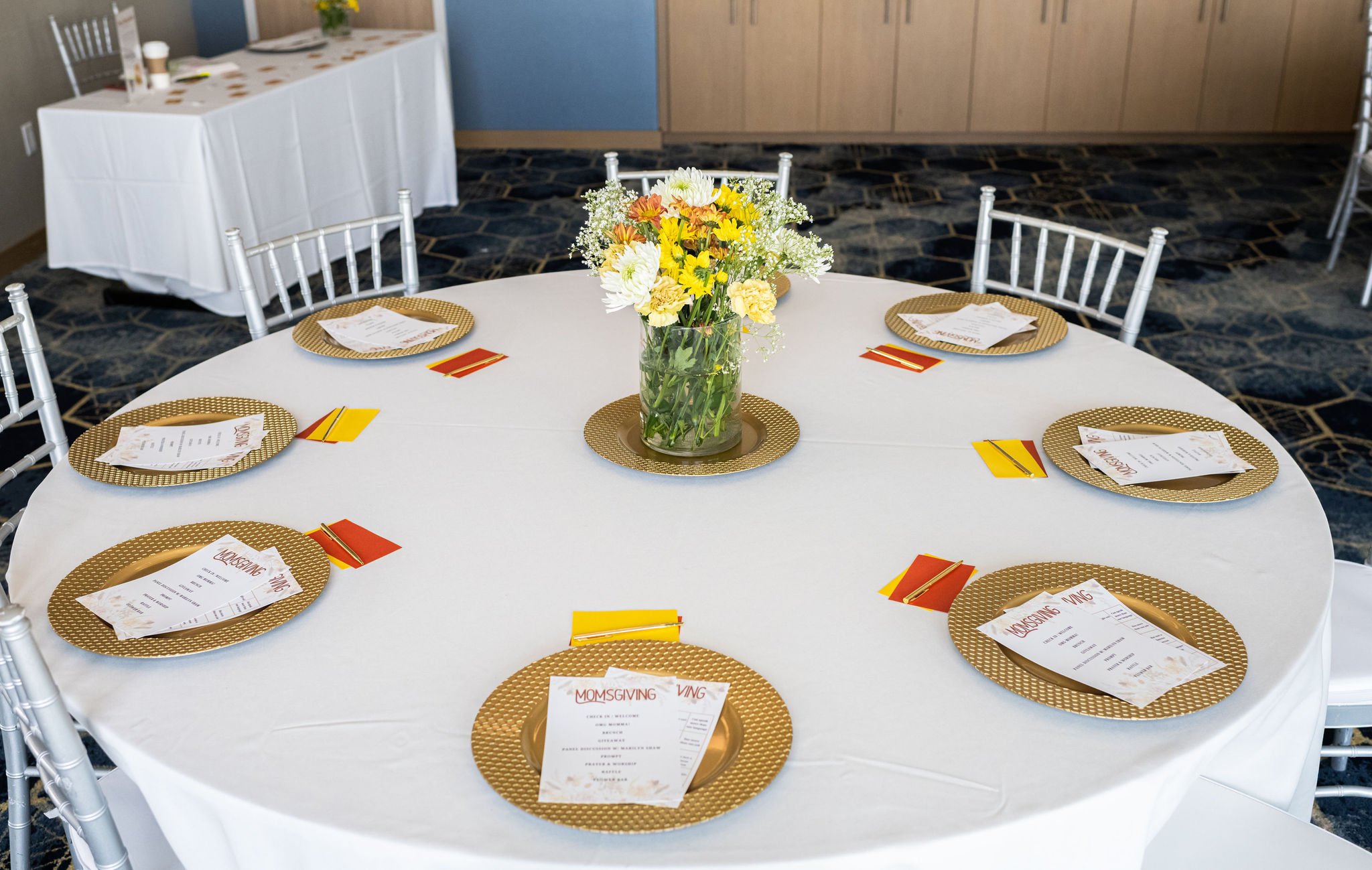 Round table with floral centerpiece, elegantly set for an event with menu cards, gold chargers, and colorful napkins.